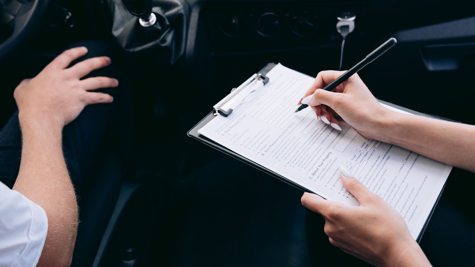 Close-up of a person writing on a clipboard inside a car, showing hands and a gear shift.