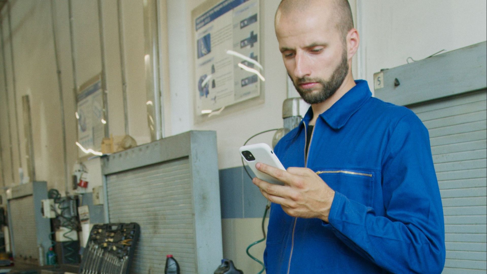 A bald mechanic in a blue jumpsuit checks his smartphone in an industrial workshop setting.