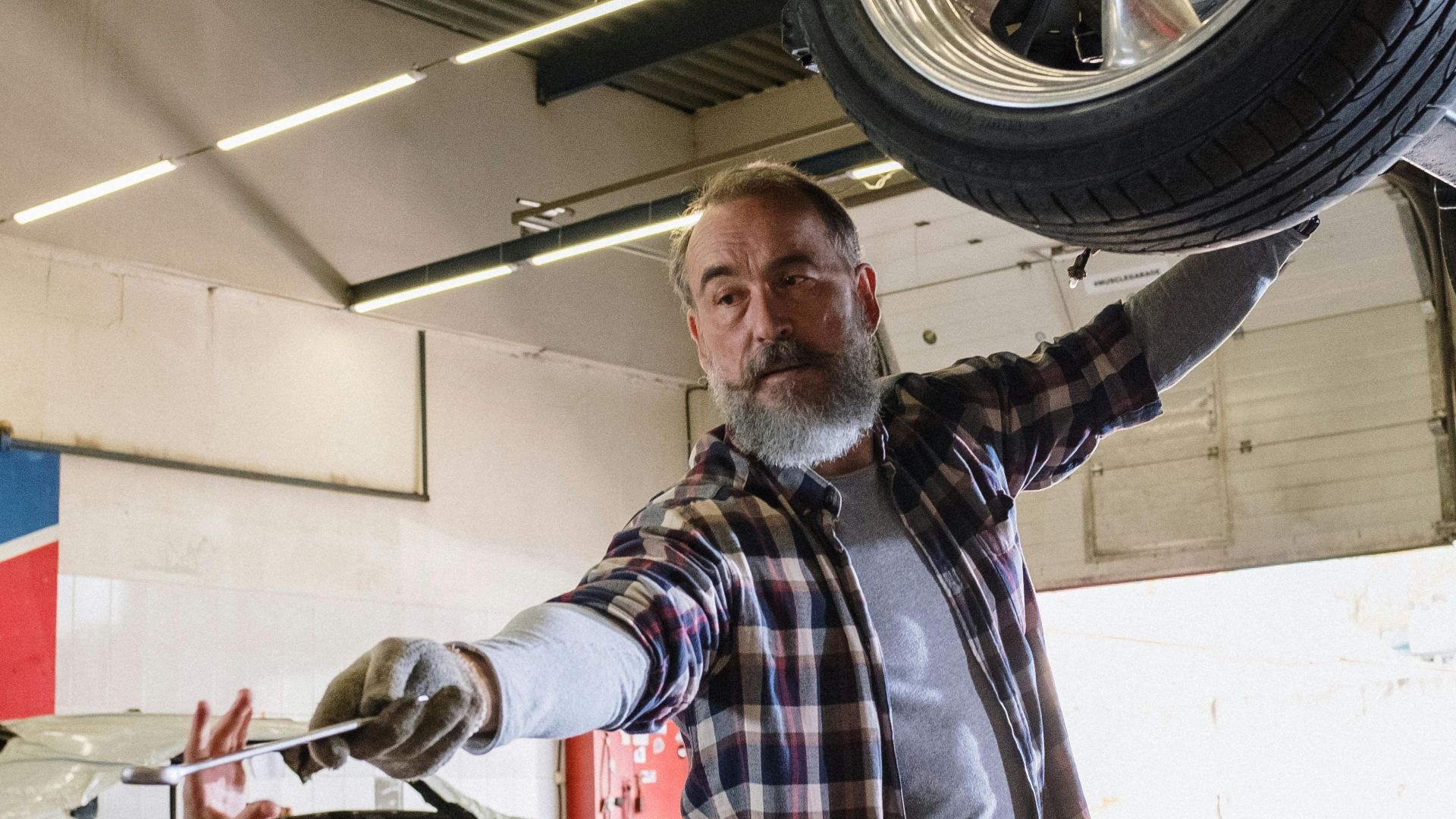 Bearded mechanic working on a vehicle in an auto repair shop, handing a tool to an assistant.