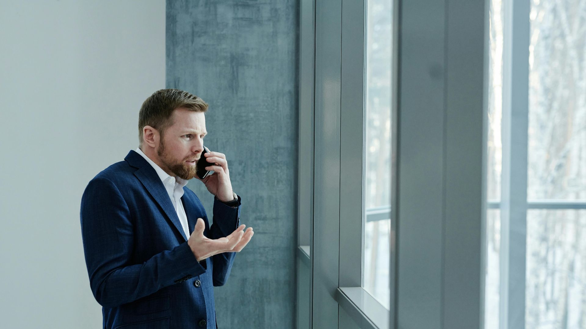 Businessman using smartphone while standing indoors by glass window, expressing concern.