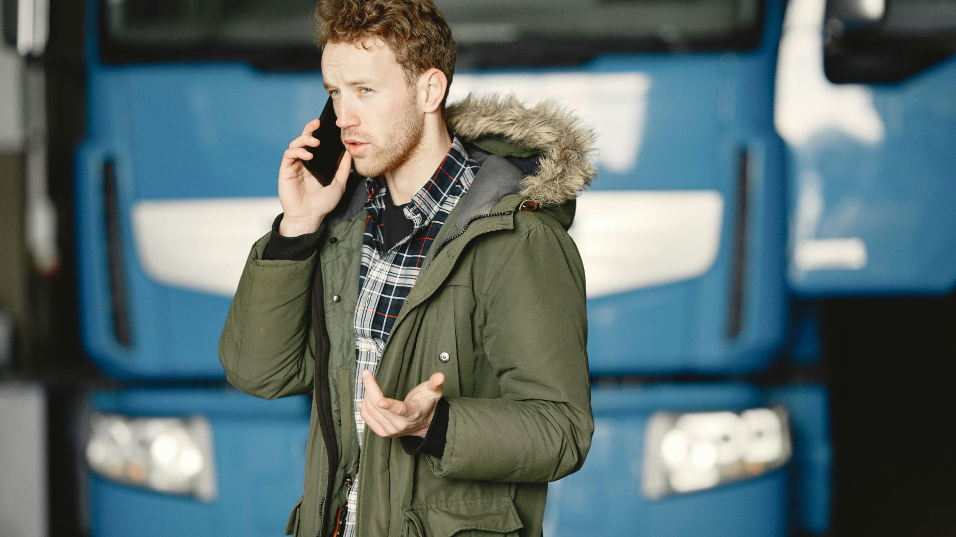 Man in a green parka talking on the phone in front of a blue truck.
