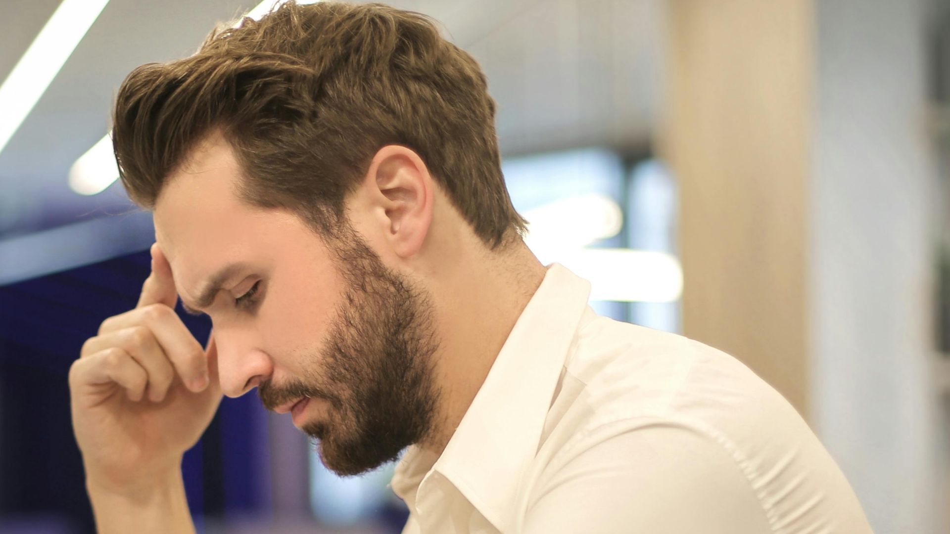 A thoughtful man in a shirt works on his laptop at a modern indoor office space.