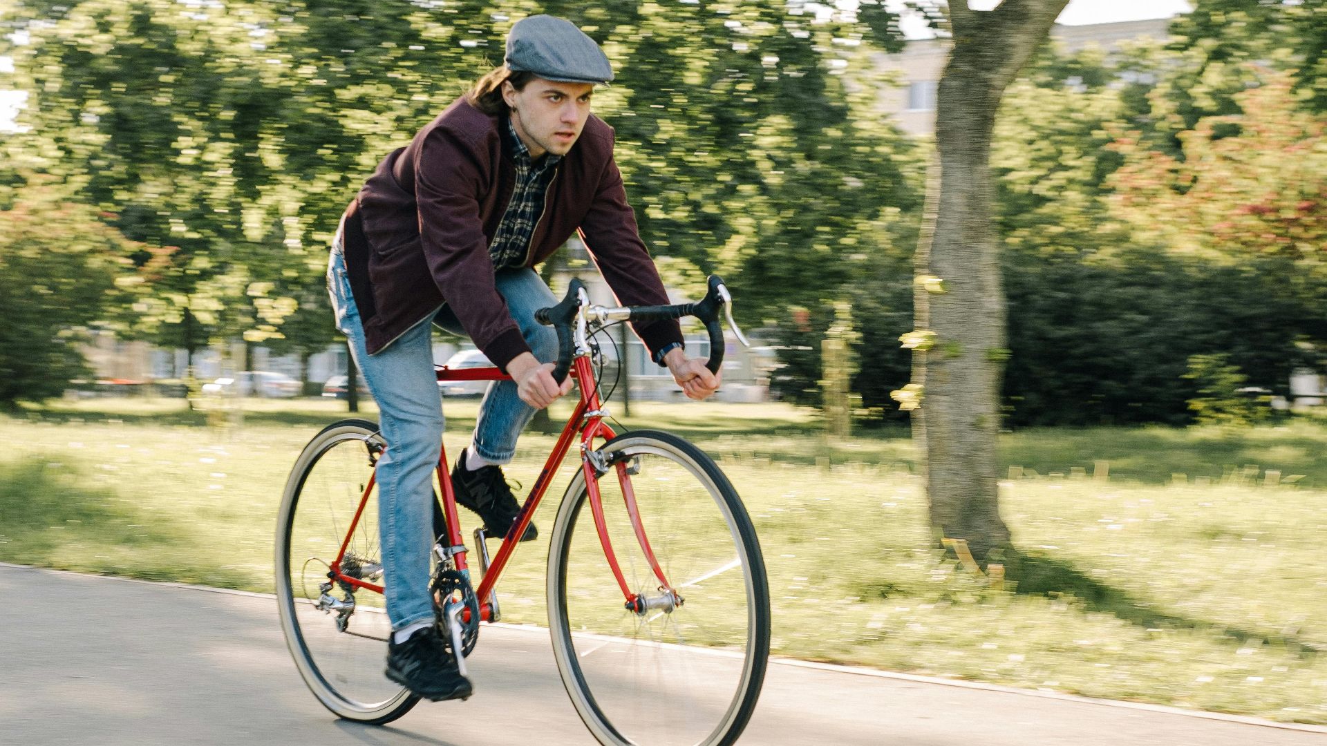 Caucasian man wearing a beret cycling on a road bike in a sunny park setting.