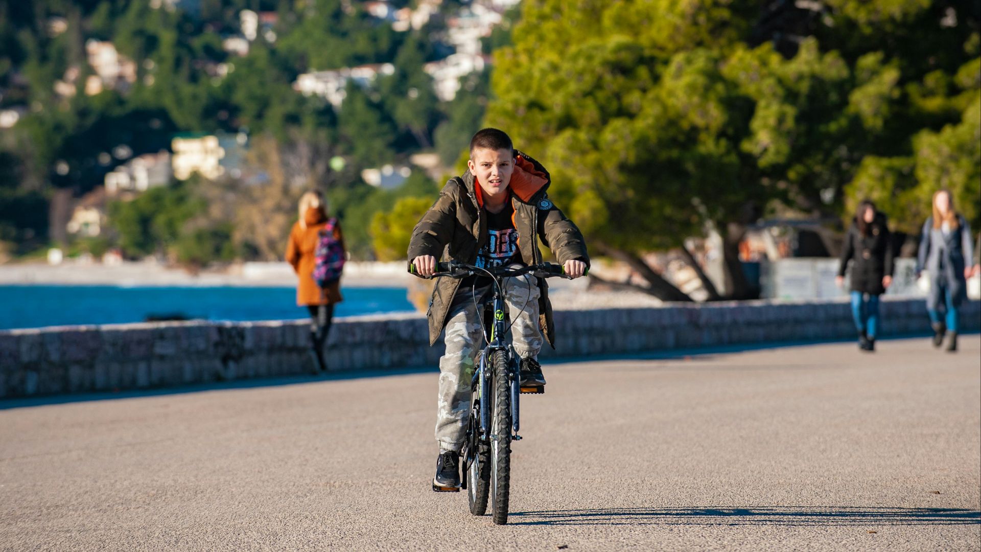 A young boy rides his bicycle along a scenic waterfront path on a sunny day.