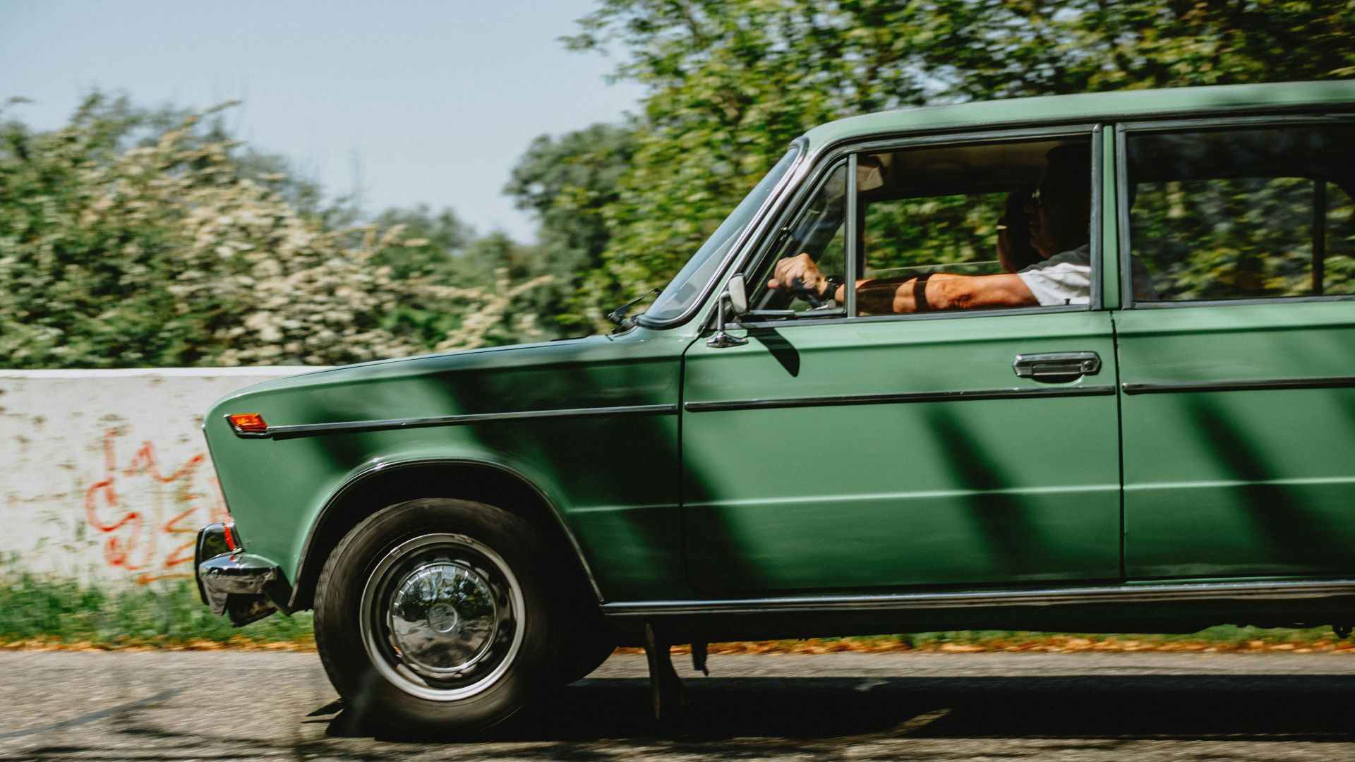 A classic green car being driven outdoors on a sunny day with a blurred background.