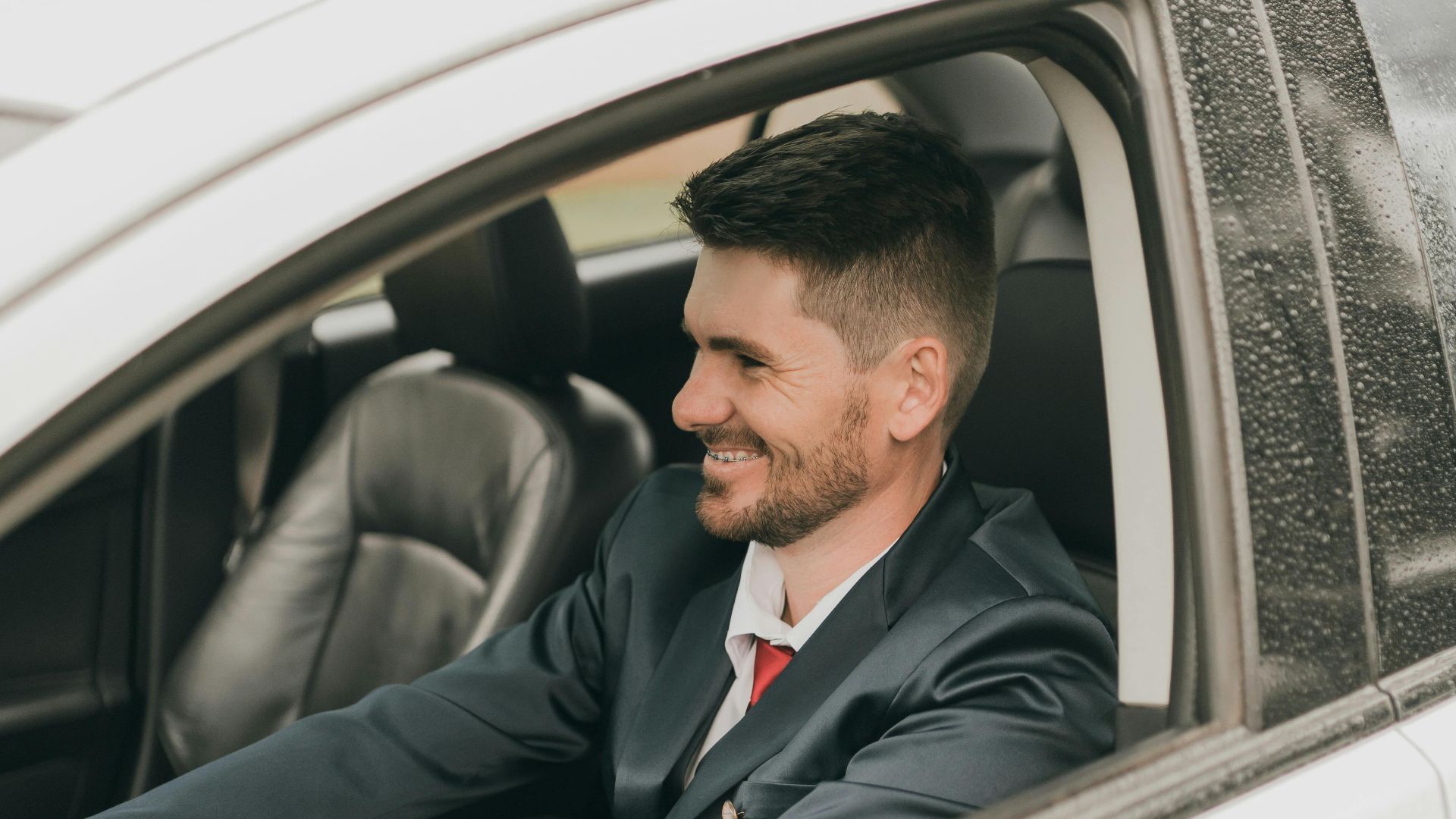 A man in a suit happily drives a silver car through city streets on a rainy day.
