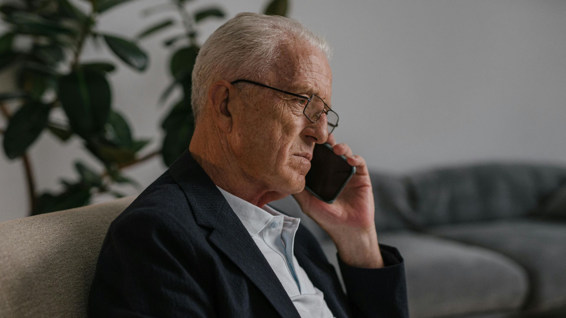 Senior man with eyeglasses sitting indoors, having a phone call. Calm and thoughtful atmosphere.