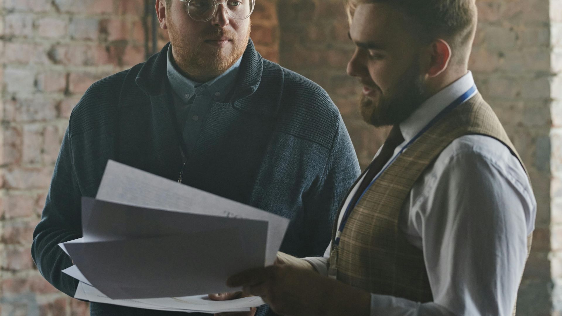 Two men discussing paperwork in a rustic office with exposed brick walls.