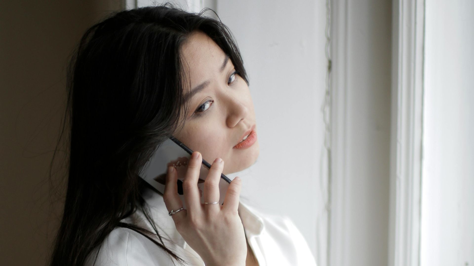 Young Asian female in casual clothes standing near window and answering phone call in cozy room at home
