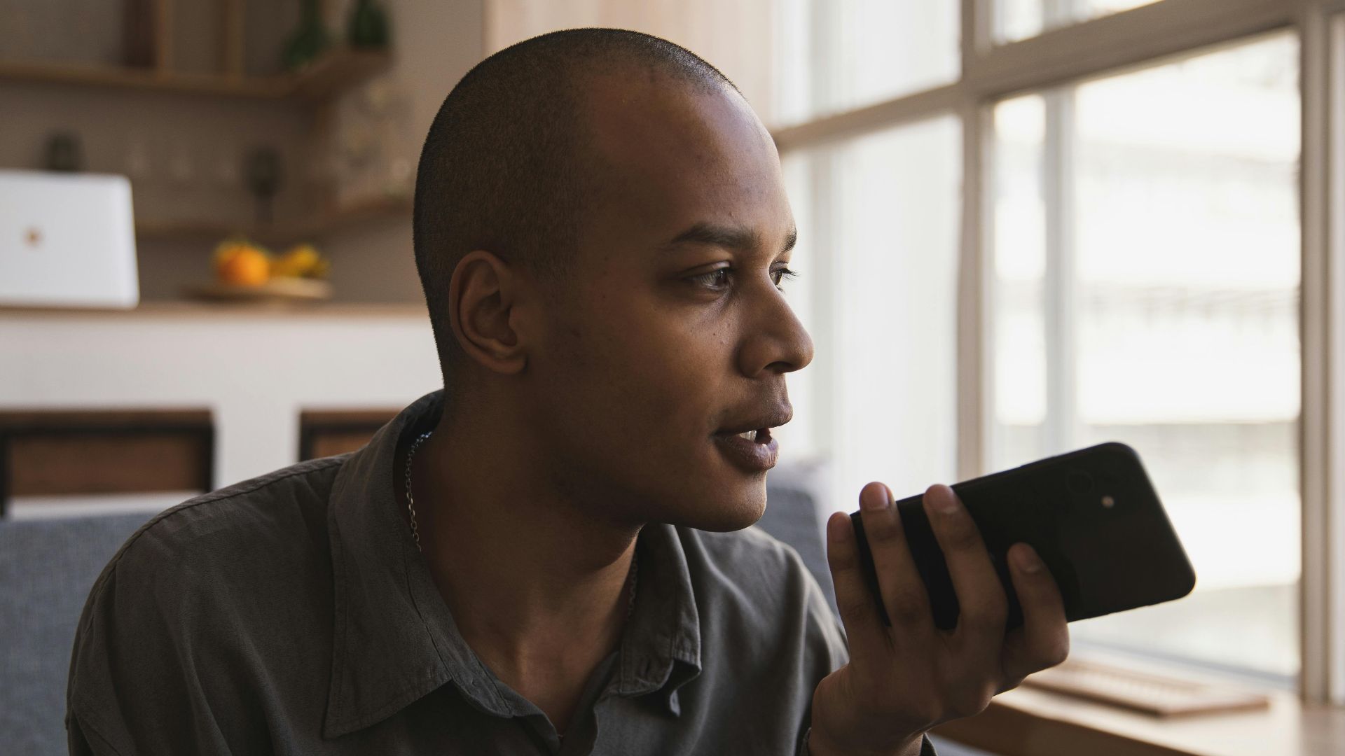 Man holding smartphone and speaking indoors, sitting on sofa by window lighting.