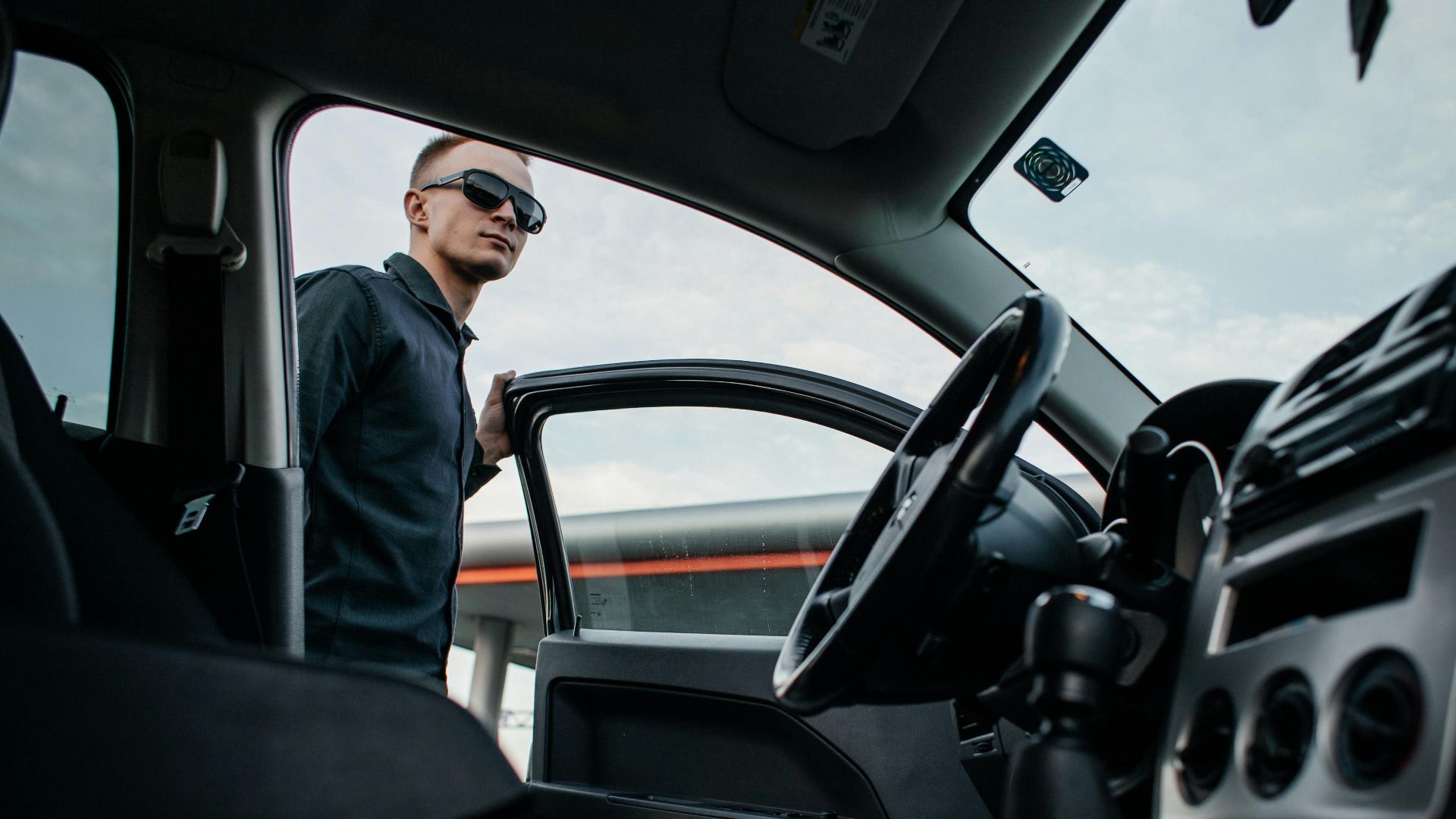A man in sunglasses stands by an open car door, viewed from the interior.