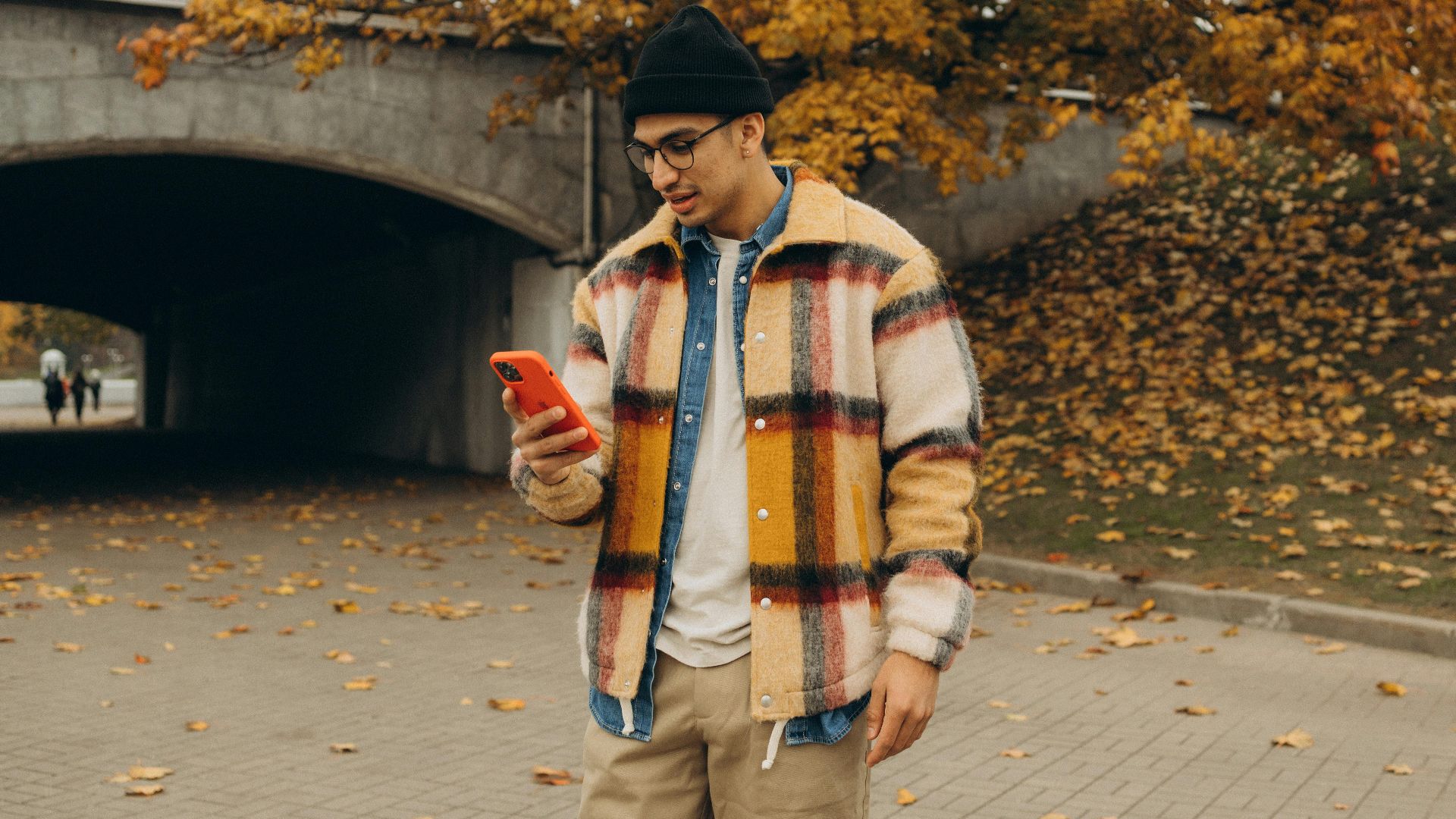 Stylish man using smartphone in park with autumn leaves and vibrant outfit.