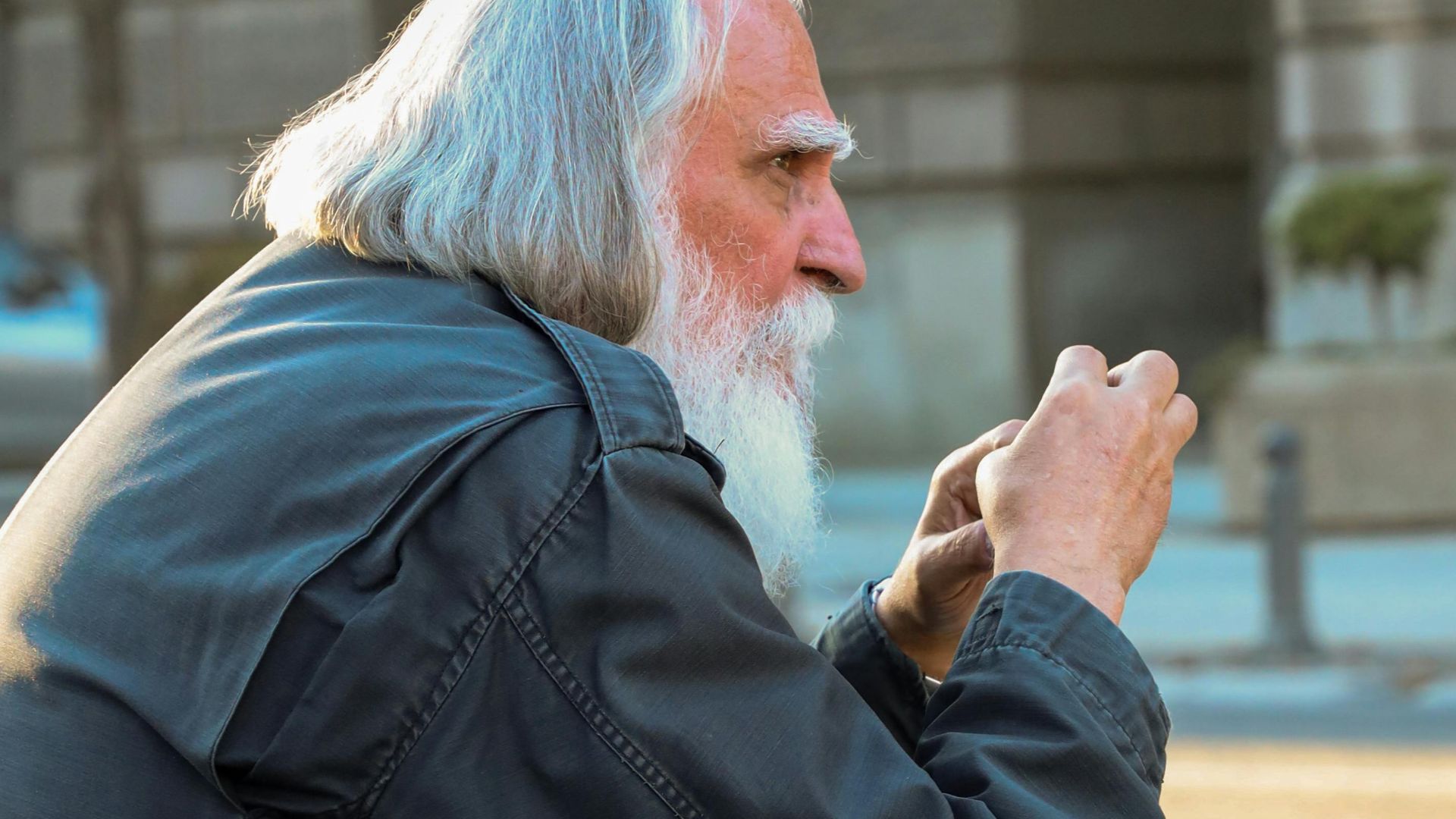 Elderly man with white hair and beard sitting outdoors in urban setting.
