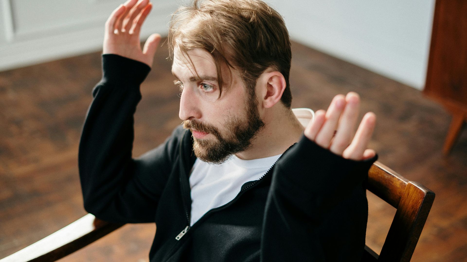Bearded man sitting and gesturing expressively in a well-lit interior space.