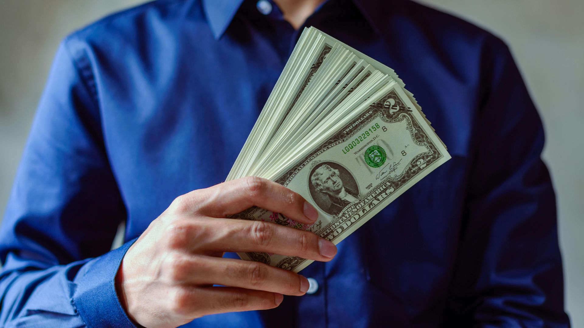 Close-up of a person in blue shirt holding multiple US dollar bills.