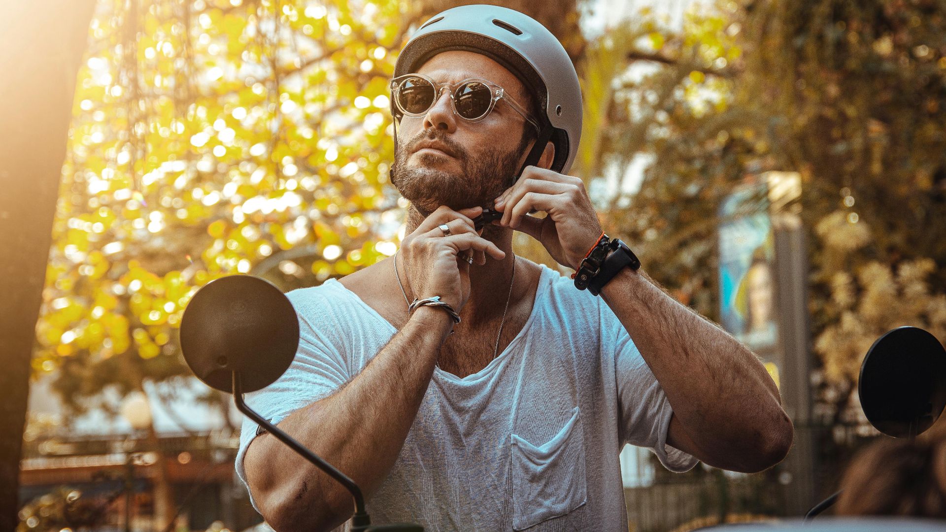 A man wearing sunglasses and a helmet adjusts his strap outdoors in bright sunlight.