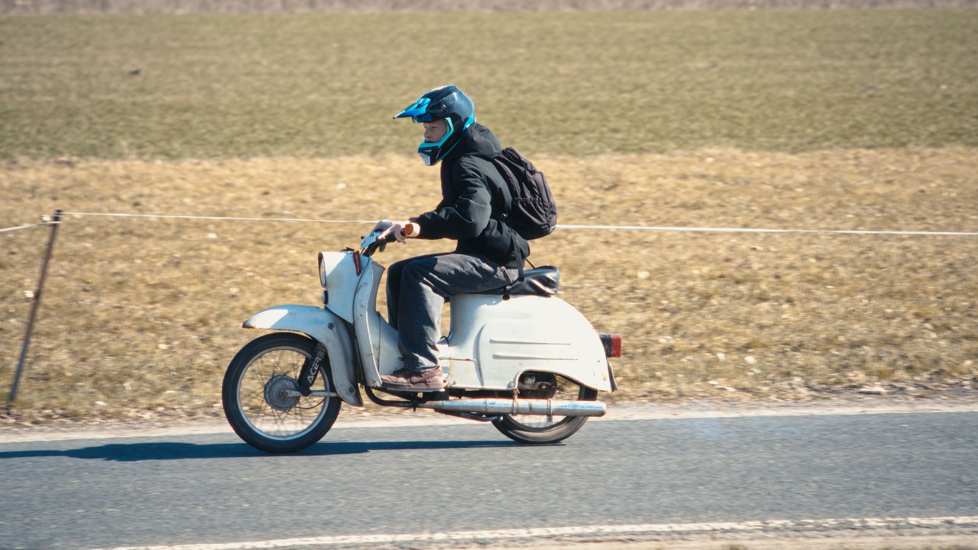 A person wearing a helmet rides a classic moped along a rural road, evoking a chill vibe.