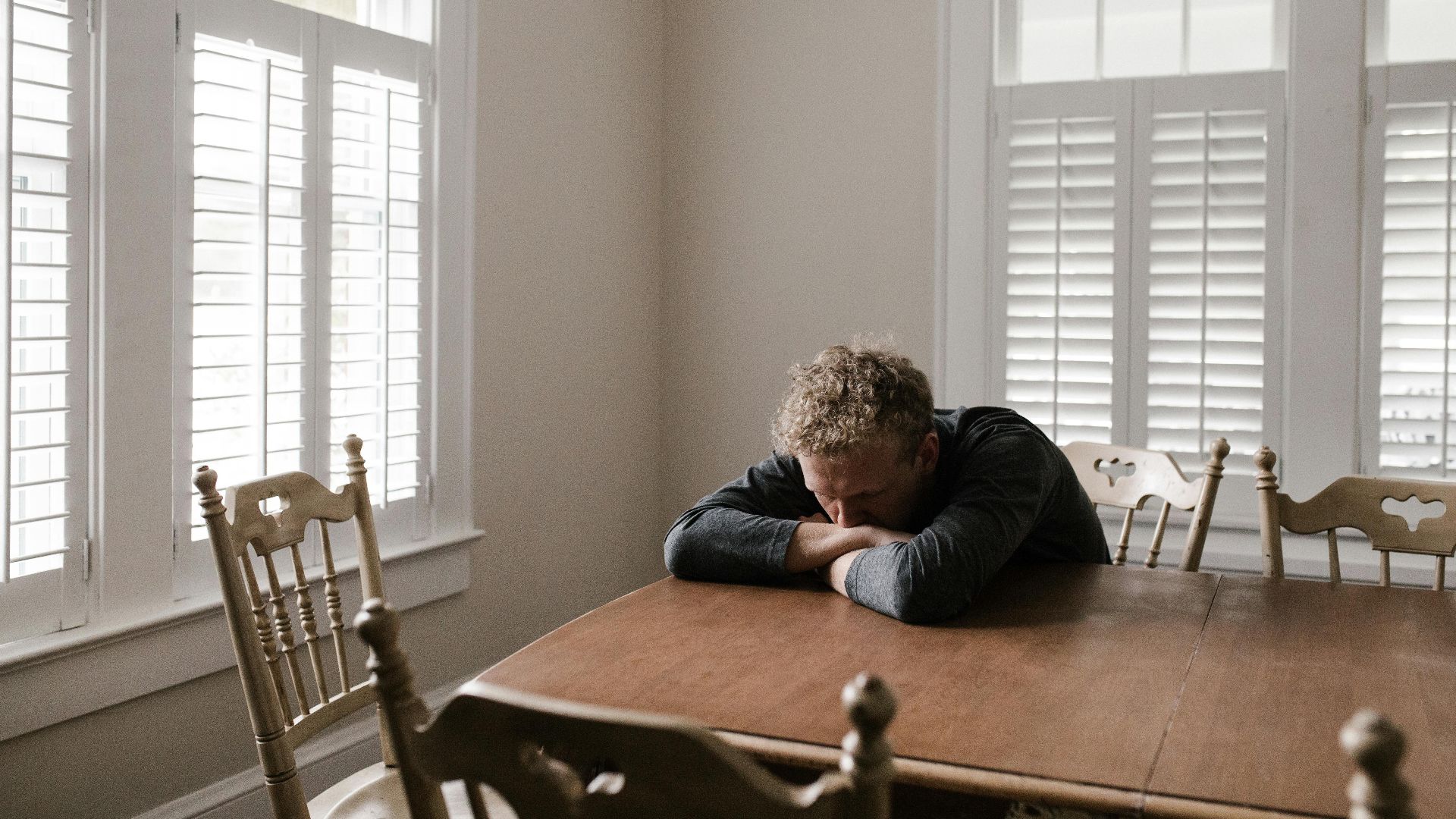 An adult man with emotions of sadness and anxiety sitting alone at a wooden table near windows.