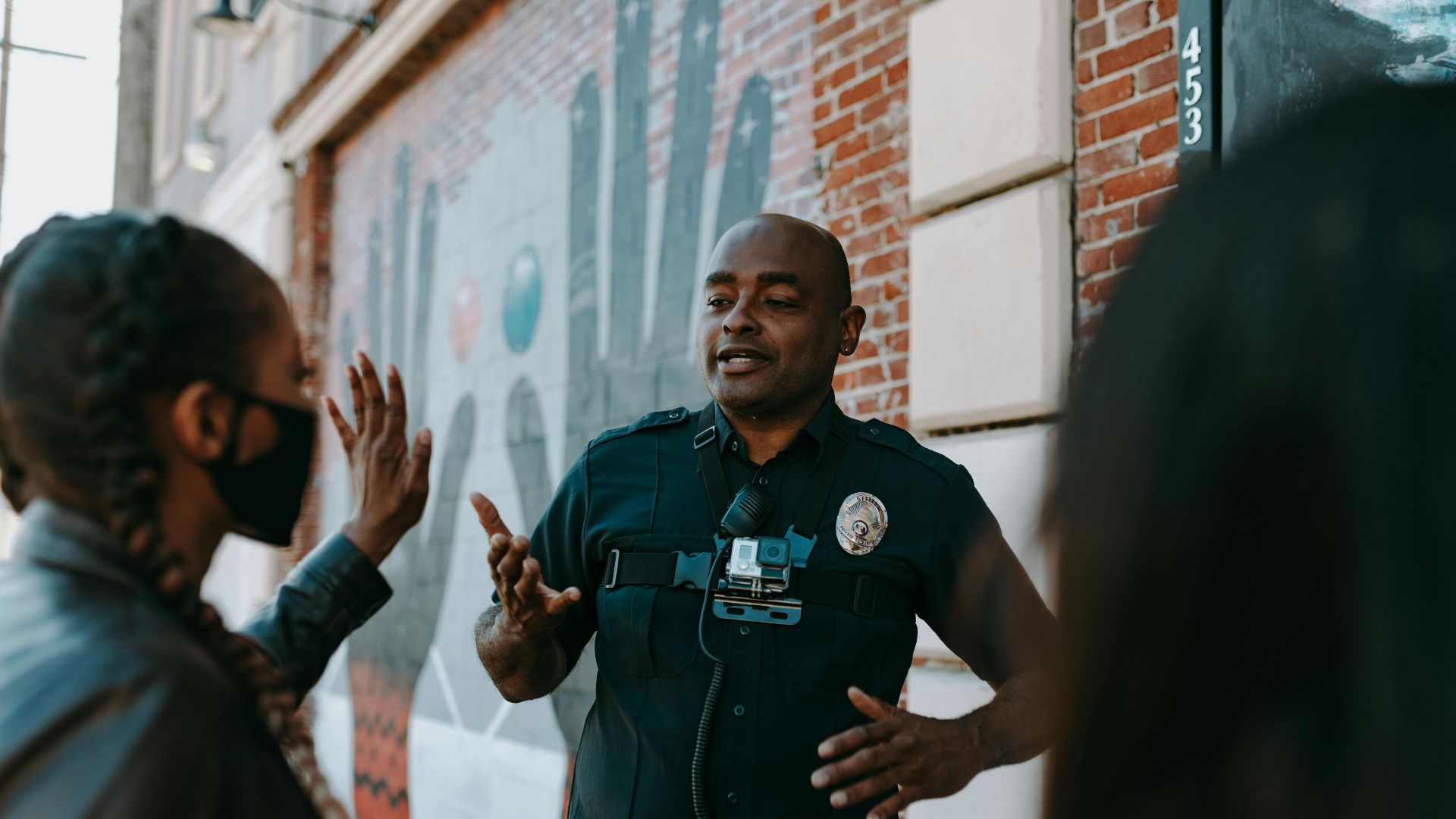 Police officer conversing with people on the street near a brick wall mural.