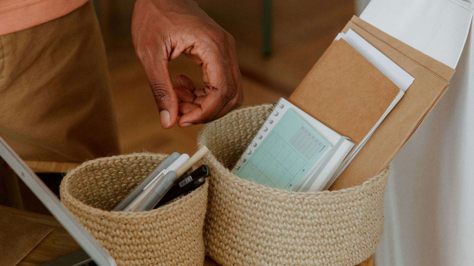 A hand reaching for stationery items in woven baskets on a desk.