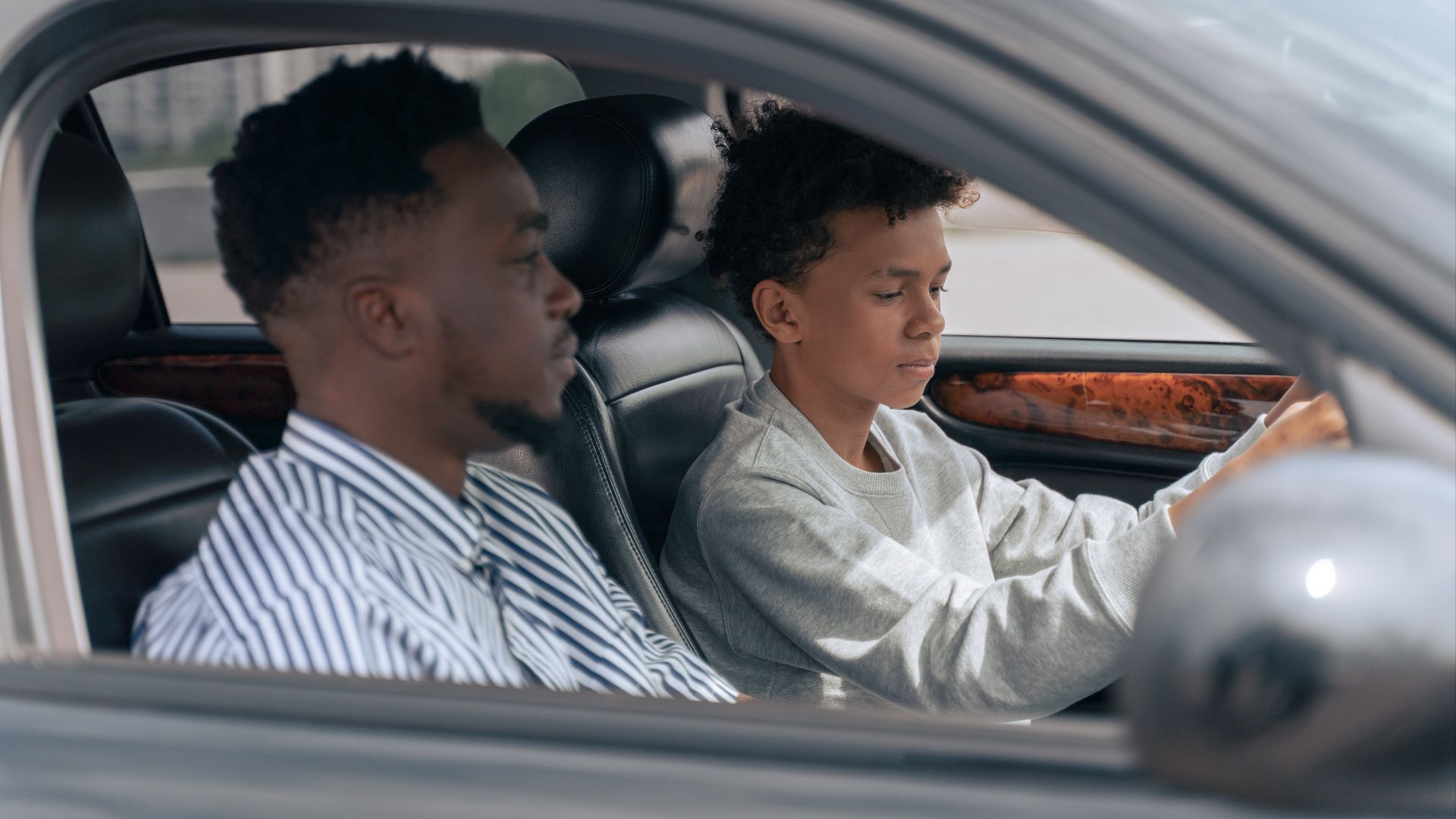 A father sits beside his son who is learning to drive, inside a car.
