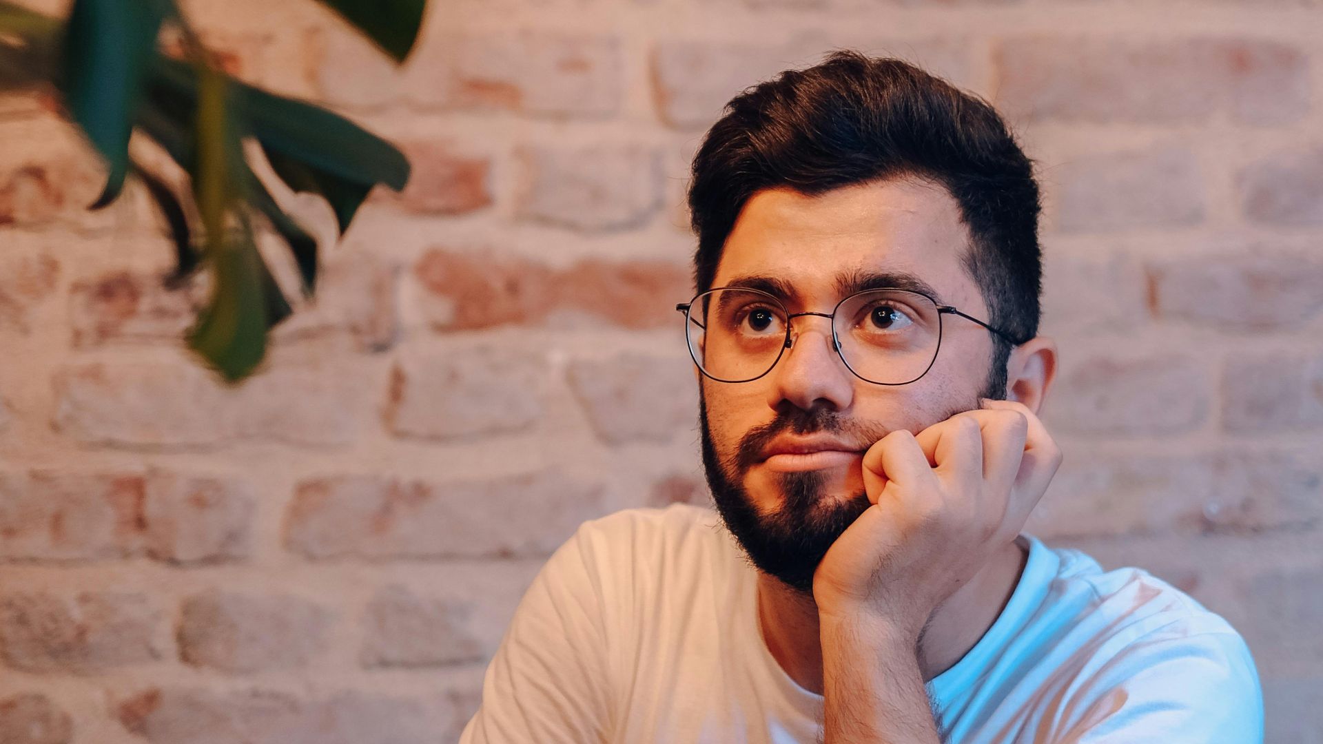 Man with glasses and beard thoughtfully looking up while seated indoors, camera on table.