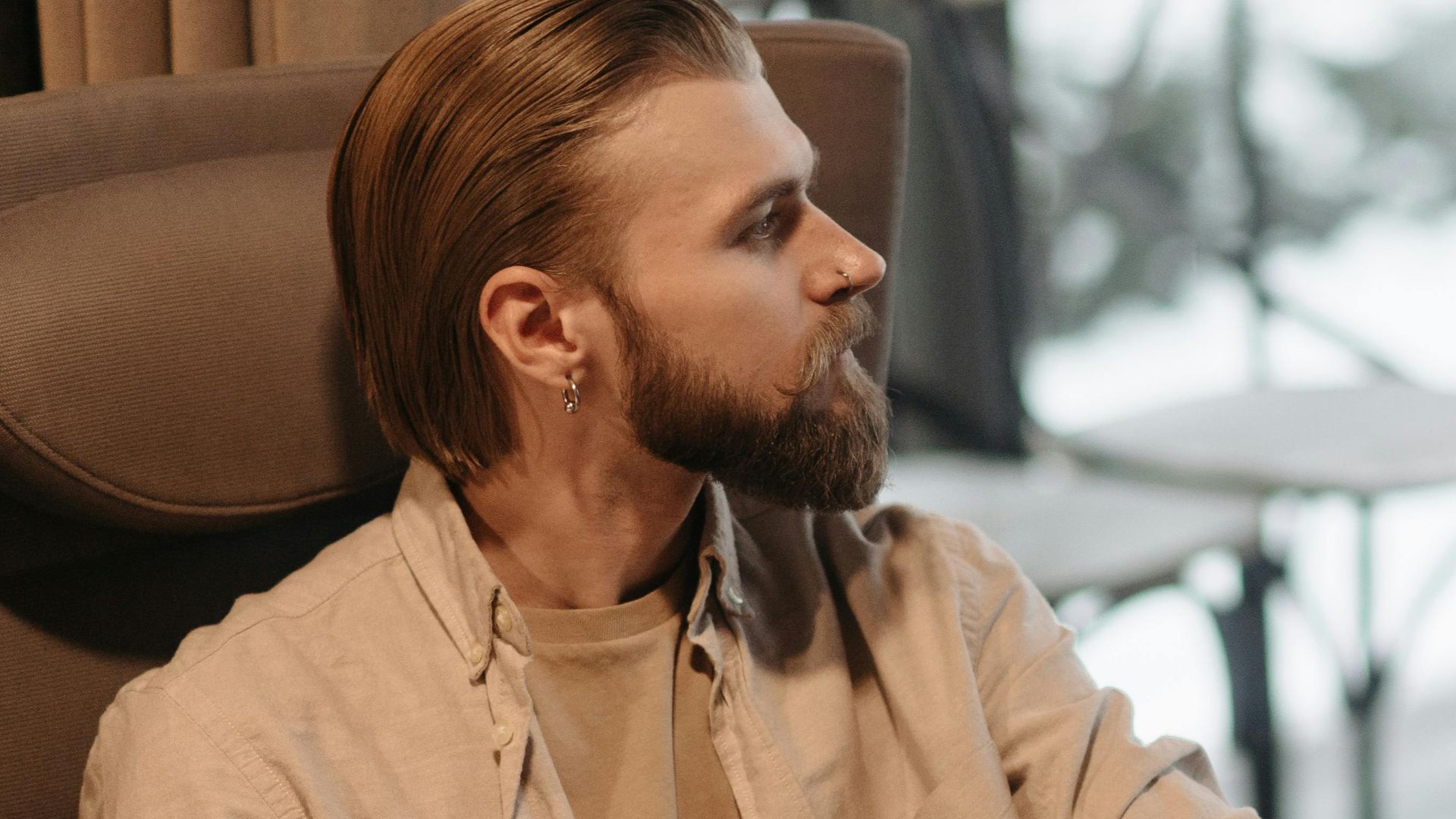 A bearded, tattooed man sits pensively, looking out a window on a winter day.