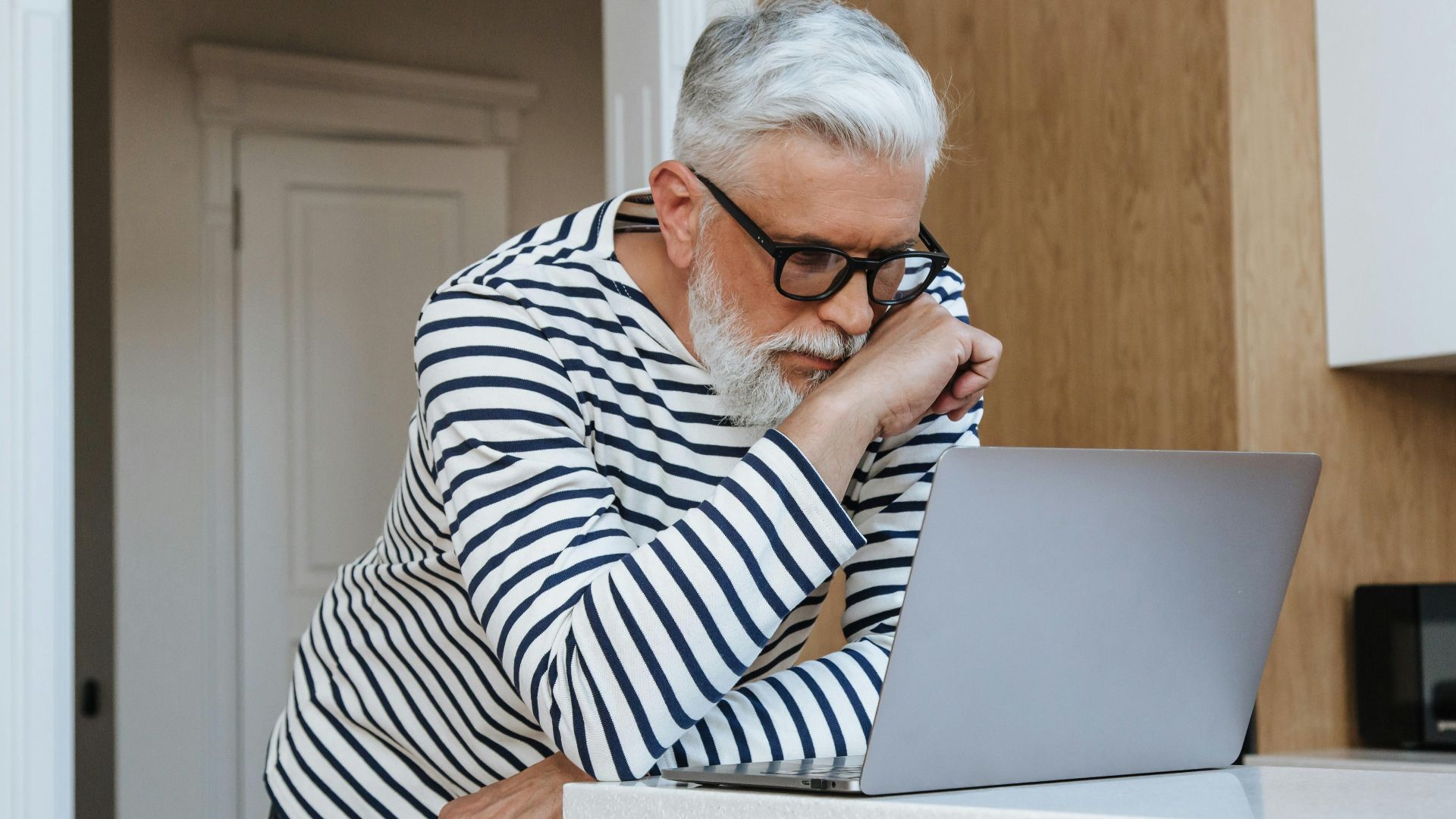 A senior adult man with glasses concentrating on his laptop at home, symbolizing modern working environments.