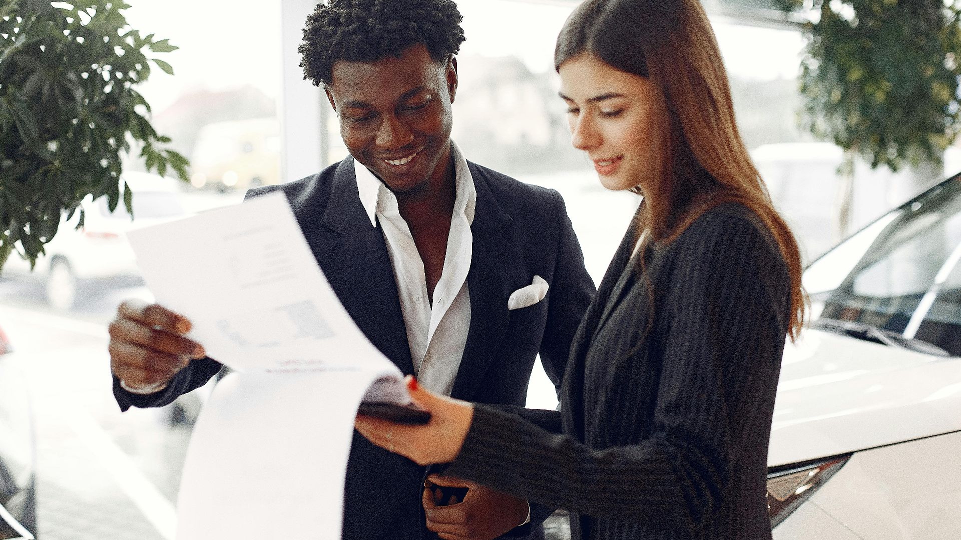 Cheerful smiling multiethnic businessman in classy suit and professional elegant female consultant standing close and reading contract details in car showroom