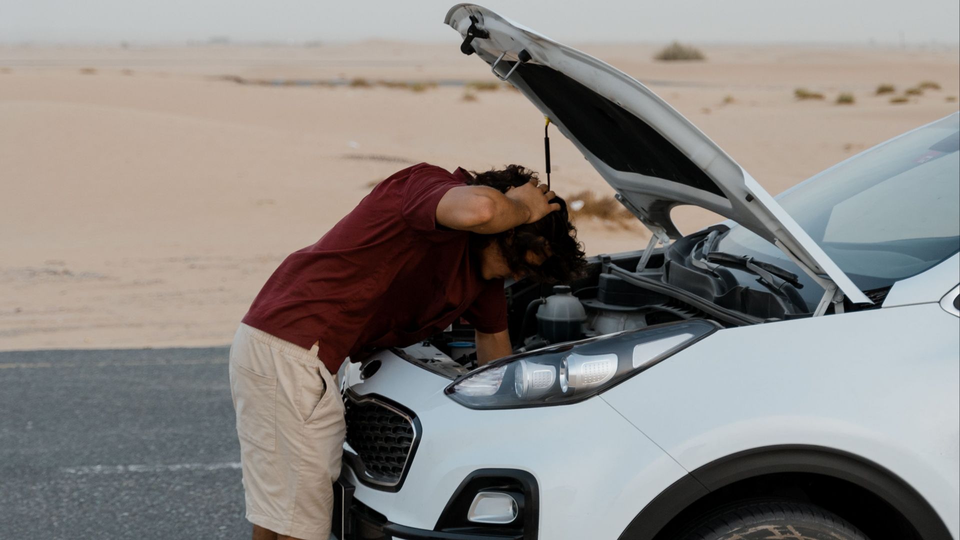 A man checks under the hood of a broken car on a deserted road, surrounded by a desert landscape.