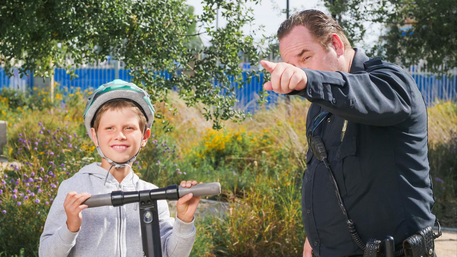 A police officer assists a young boy on a scooter at a park, signifying community safety.