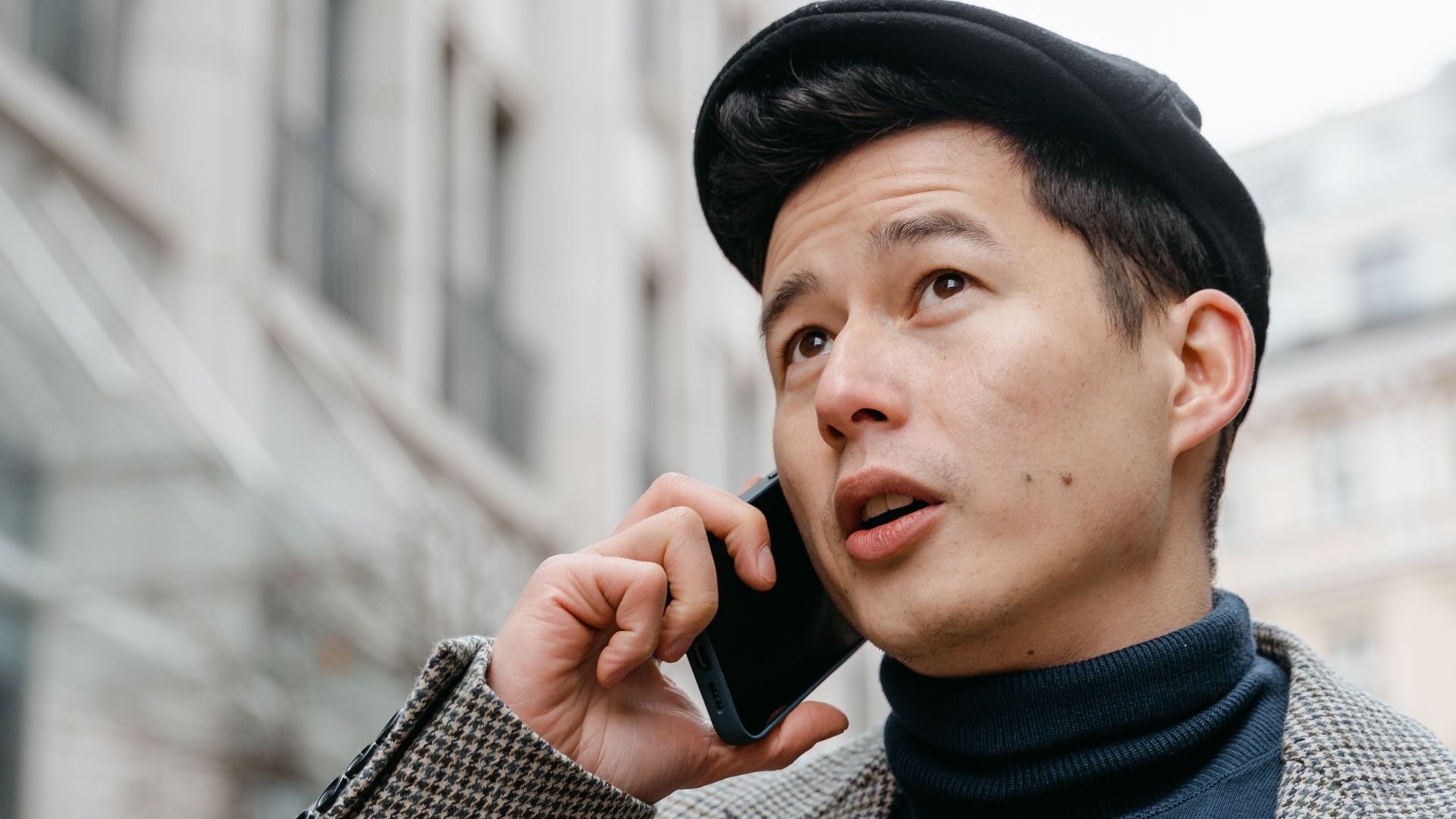 Stylish man in a city talking on a smartphone outdoors, wearing a coat and hat.