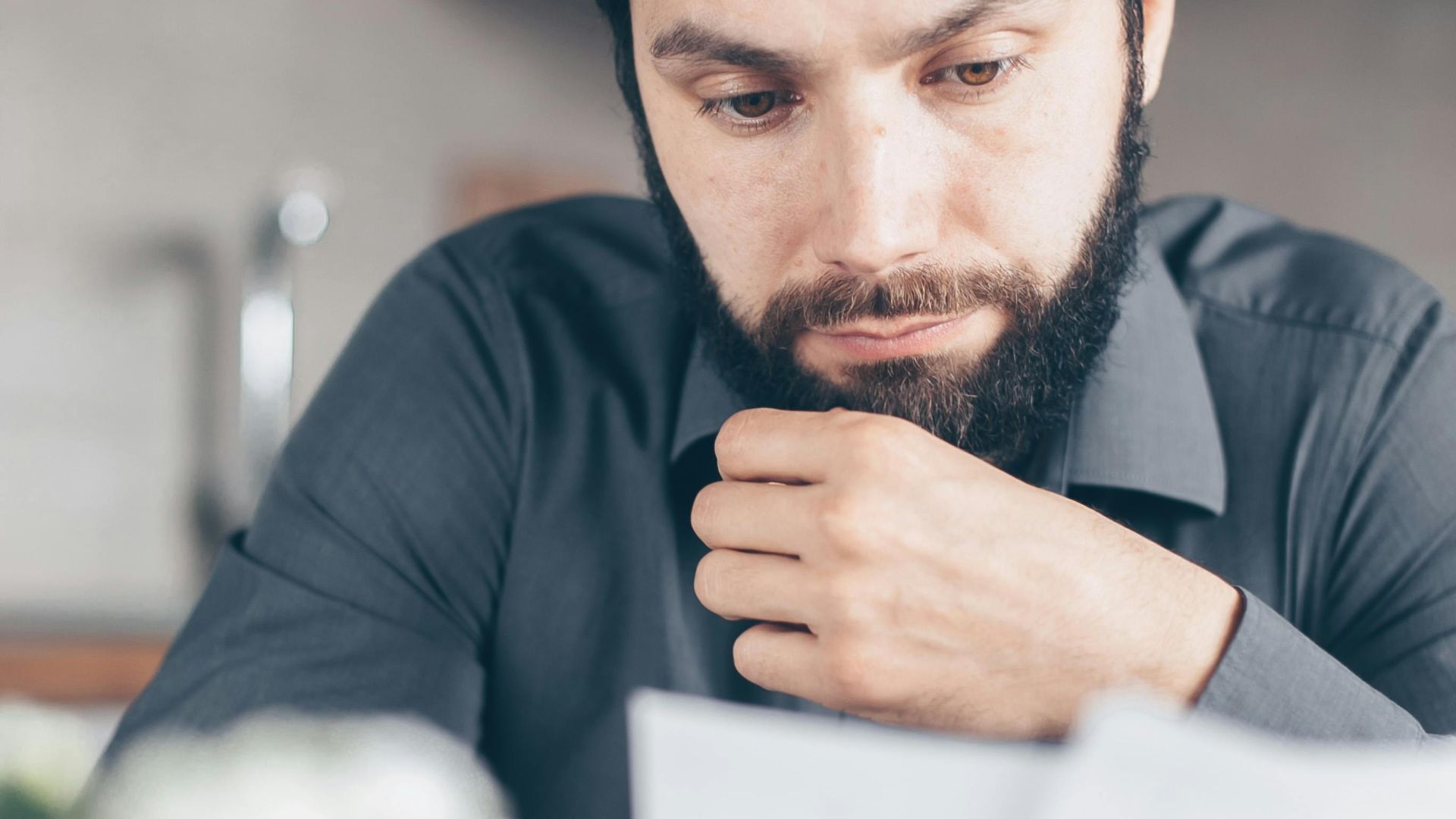 A bearded man in a gray shirt intently reads documents while seated indoors, appearing concerned.