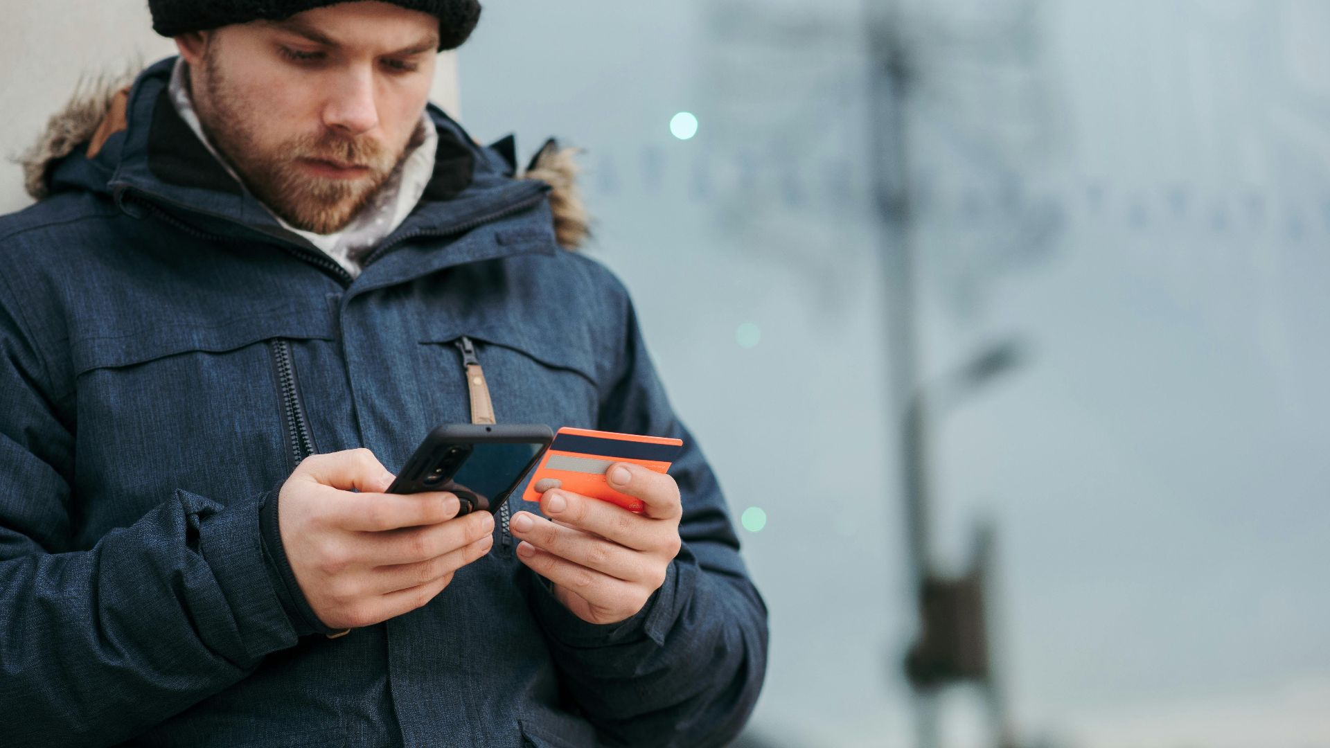 Crop concentrated man in warm clothes entering credentials of credit card on mobile phone while standing in street in daytime