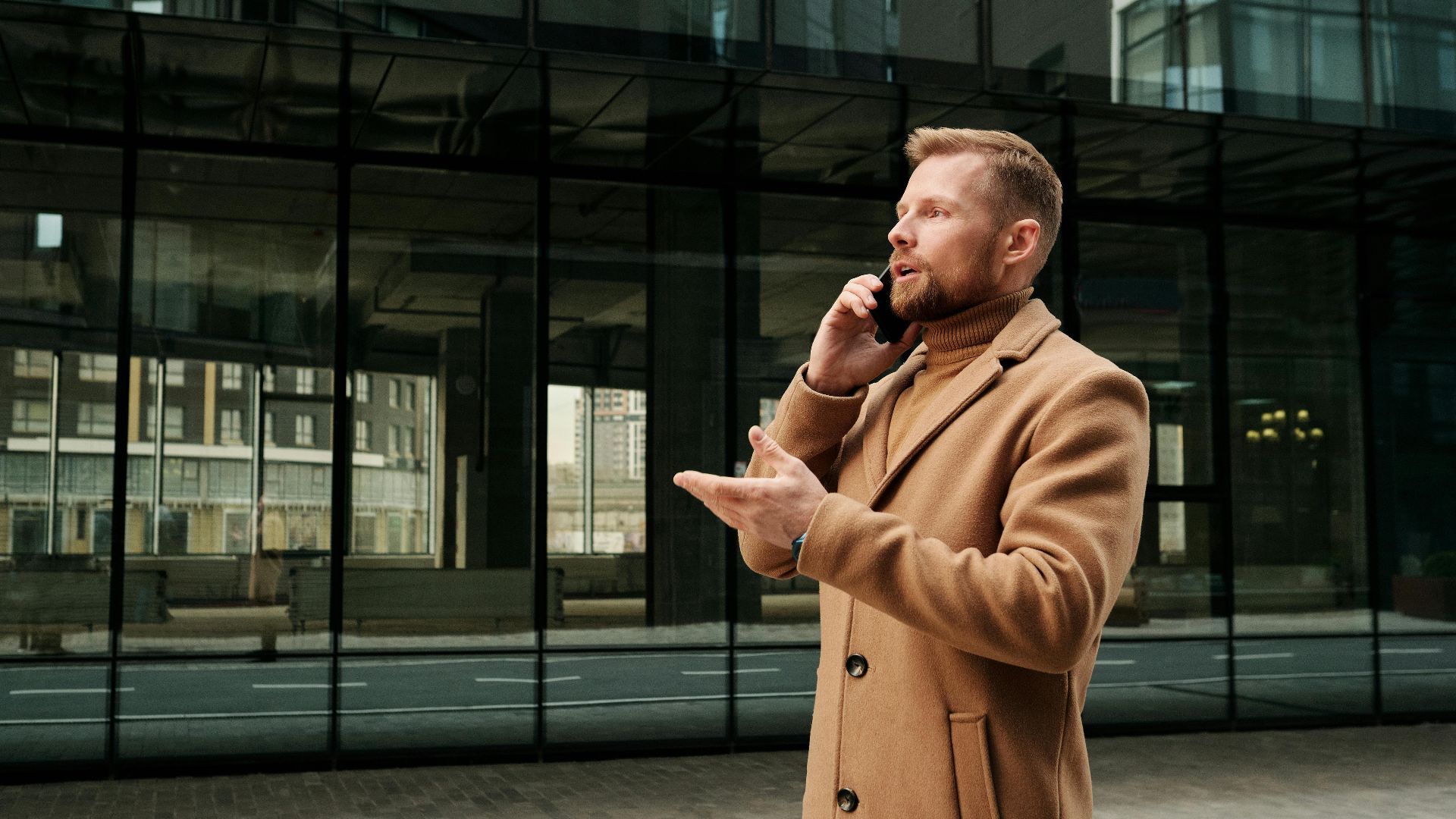 Confident businessman in a brown coat talks on phone outdoors in city.