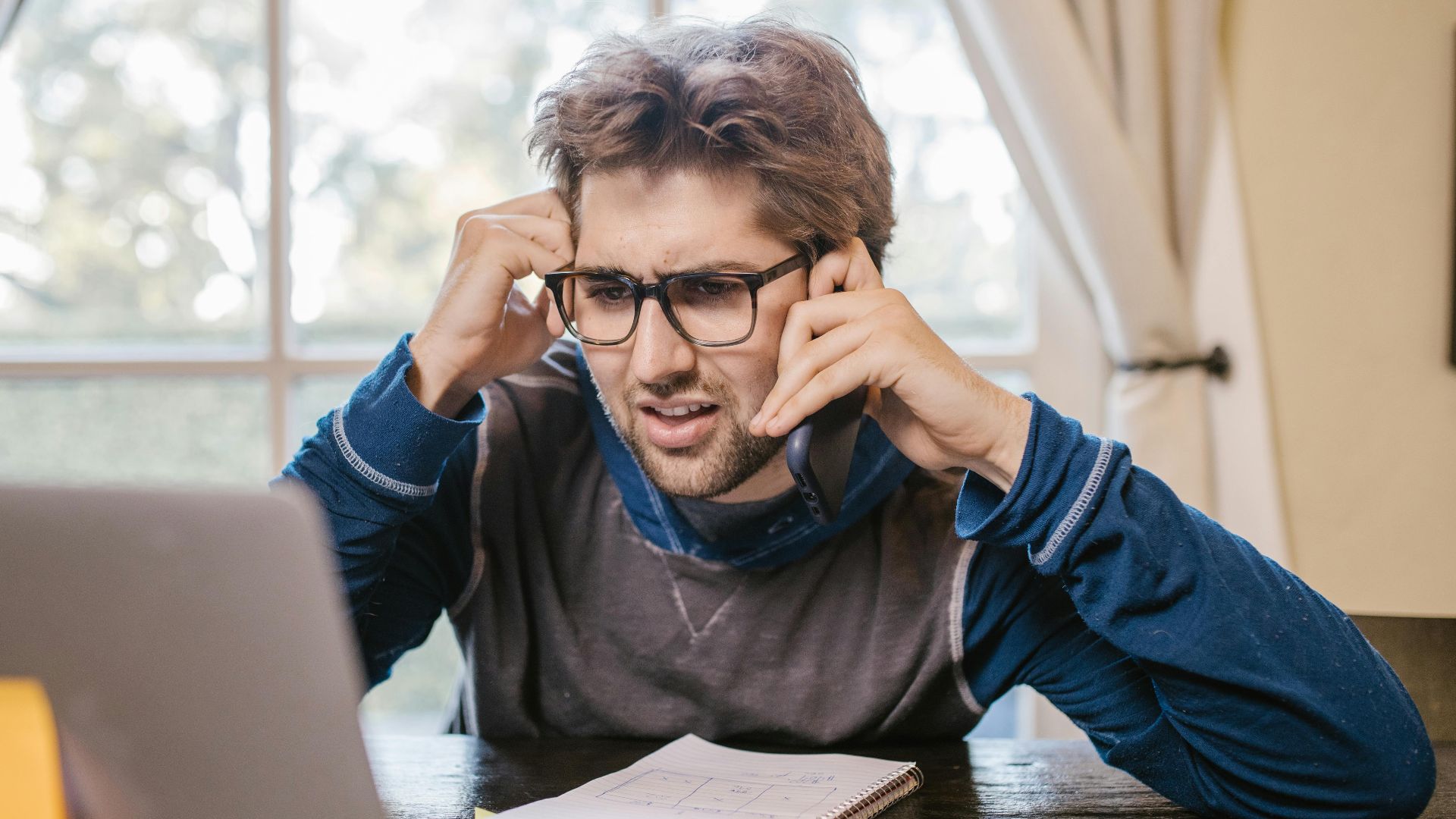 Man in glasses using laptop and smartphone at home, appearing confused.