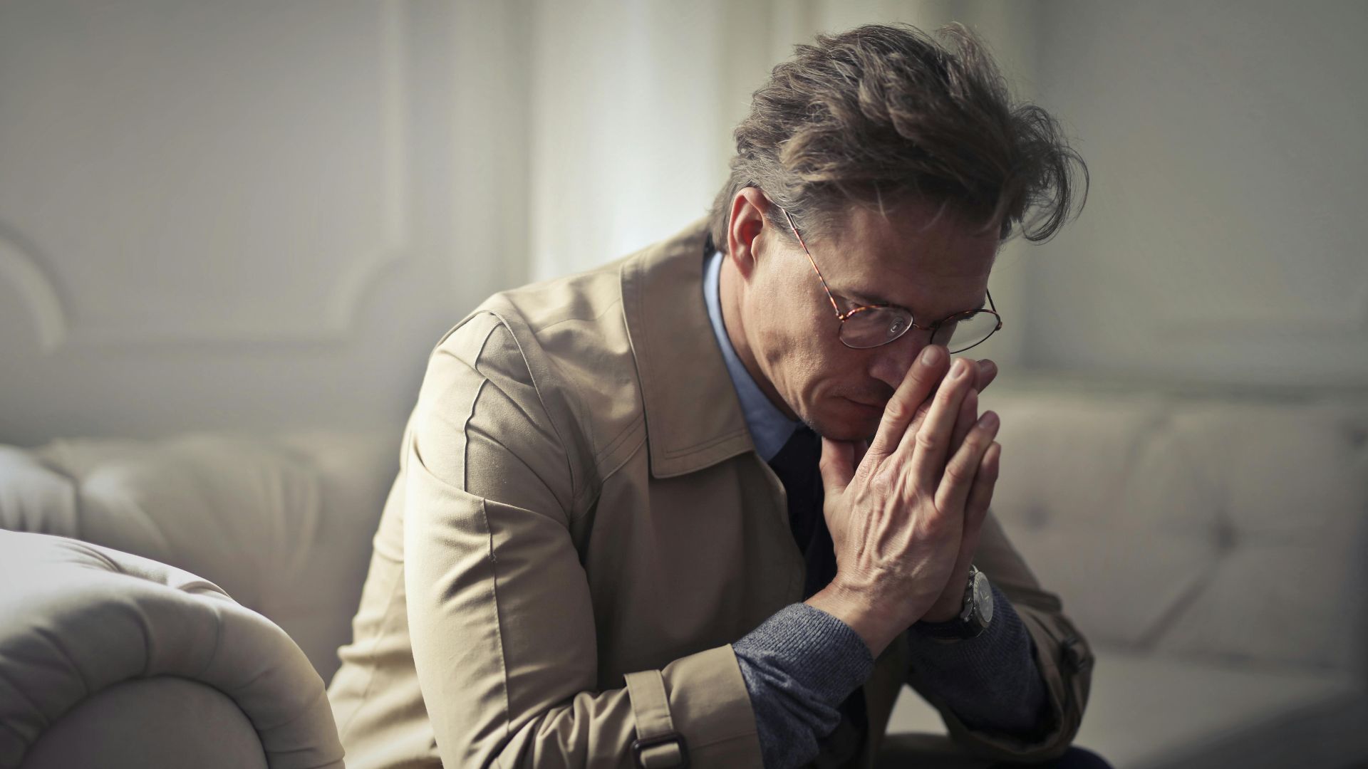 Side view of upset male entrepreneur in formal wear sitting on couch in modern living room and contemplating about financial problems while leaning on hands and looking down
