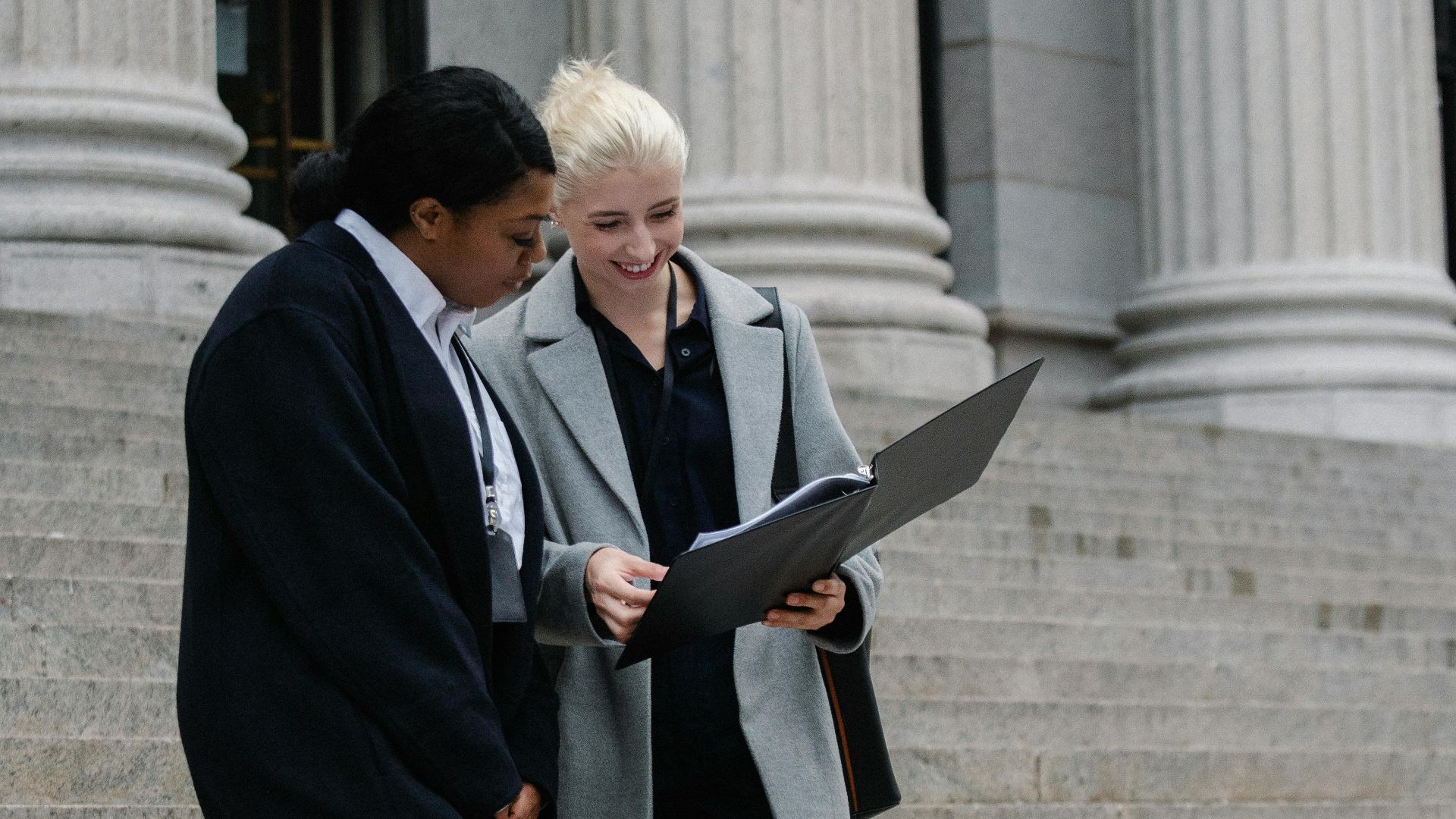 Full length of positive young multiracial female lawyers in formal outfits reading documents while standing on stairs of aged columned courthouse