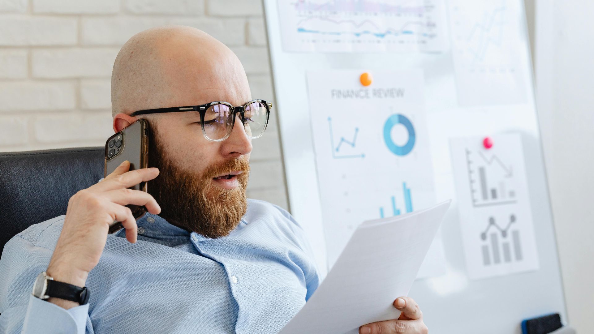 Bald man with beard holding smartphone and papers, reviewing financial charts in office.