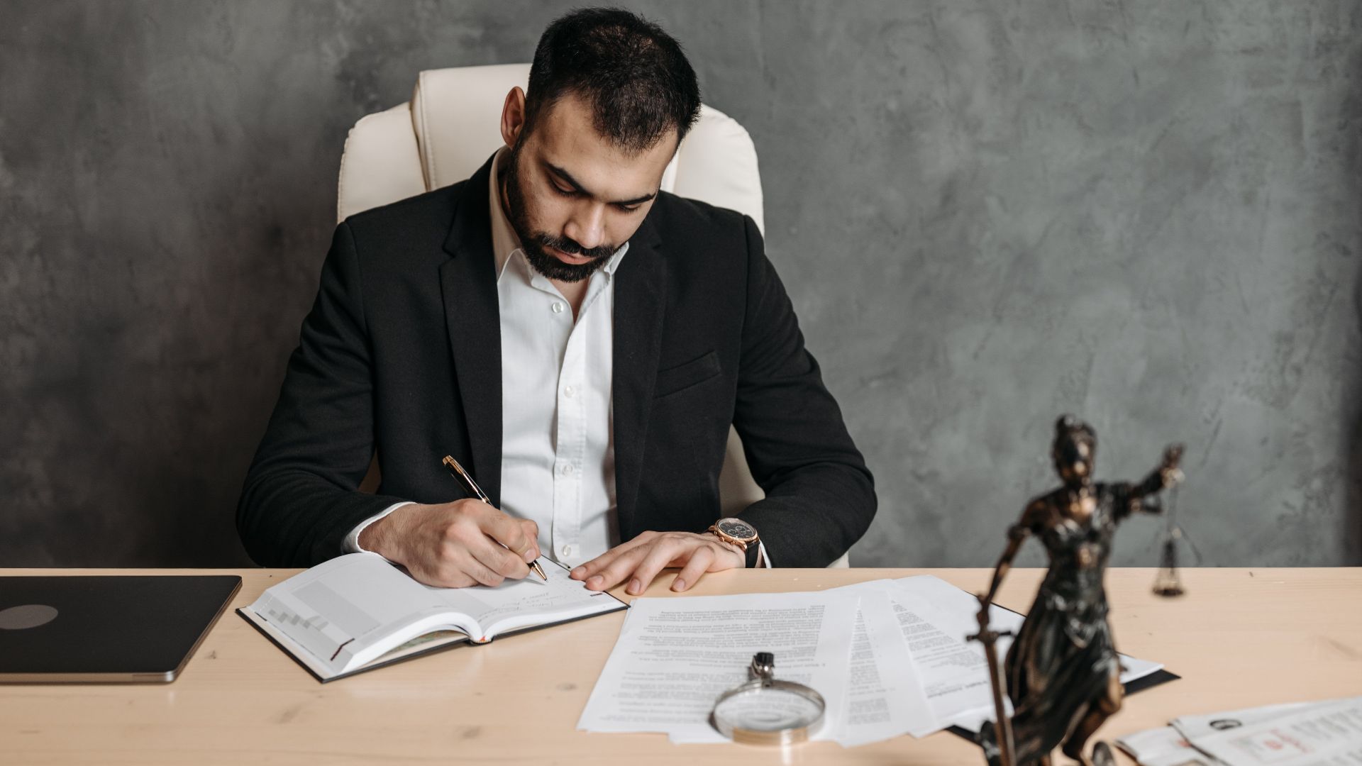 Focused lawyer in black suit at desk writing on documents in an office setting with legal statue.