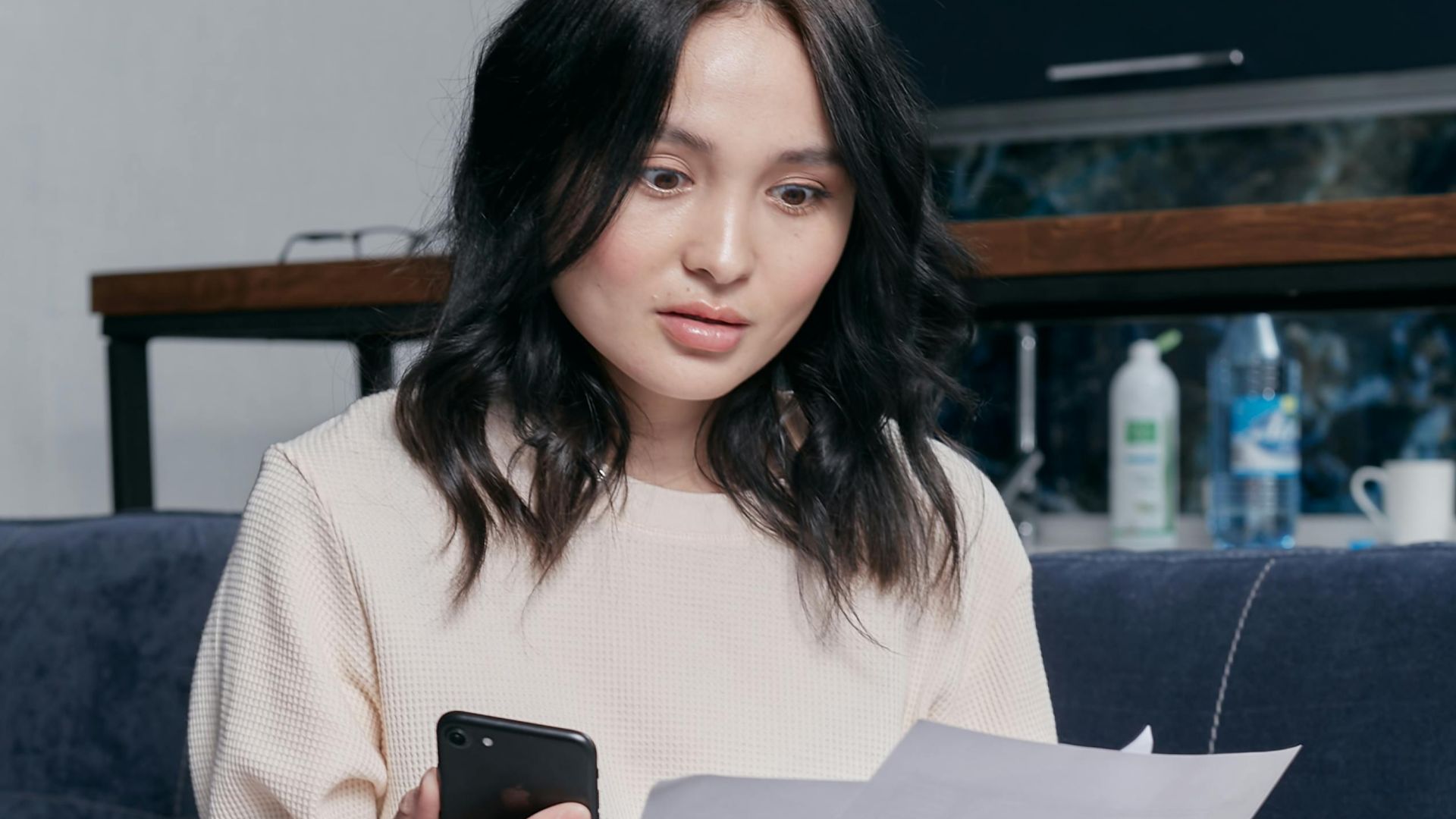 Young woman reviews documents and phone indoors, showcasing concern while seated on a couch with scattered papers. Business concept.
