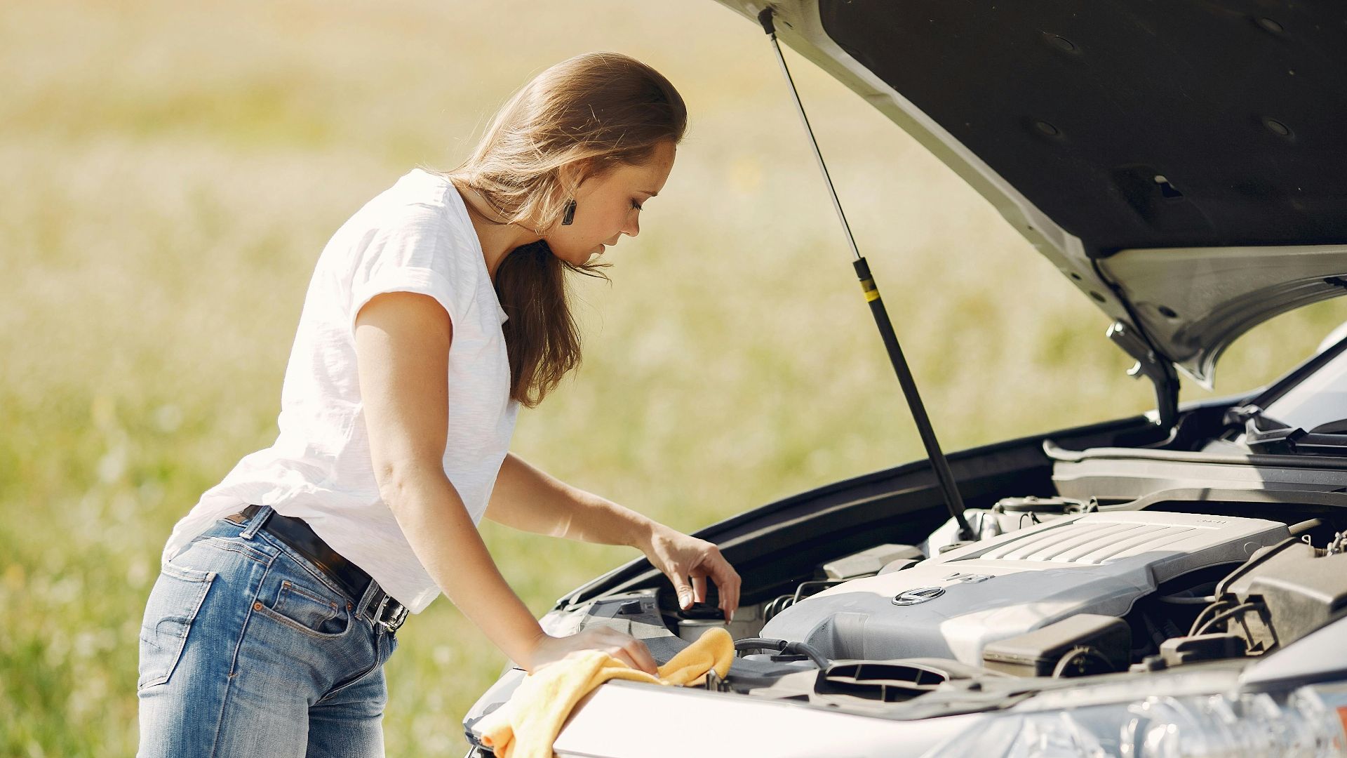 Side view of female driver in white t shirt and jeans checking damage of car engine and wiping details under open hood while standing against green field in summer day