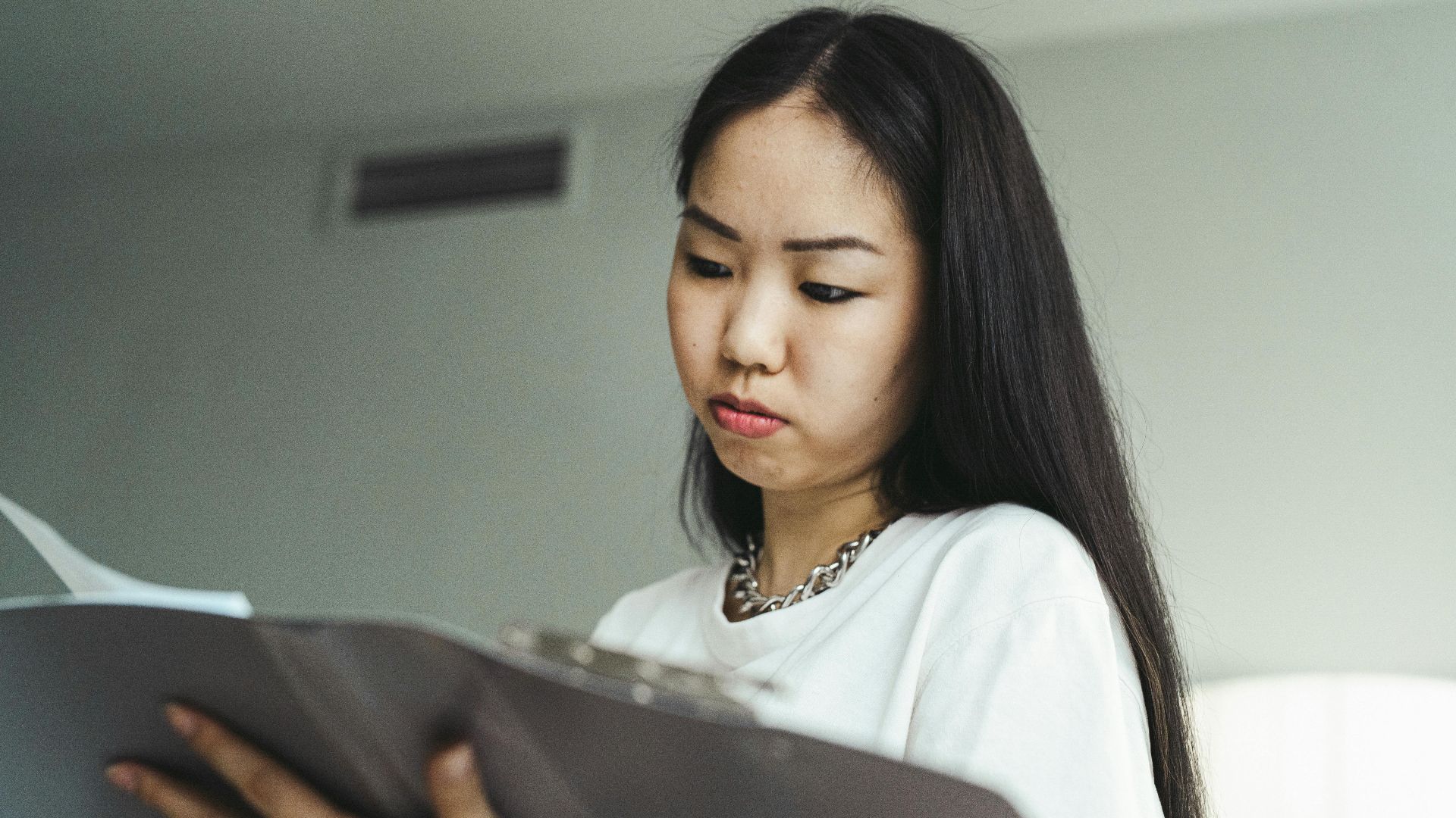 Asian woman in casual attire examining paperwork with concentration indoors.