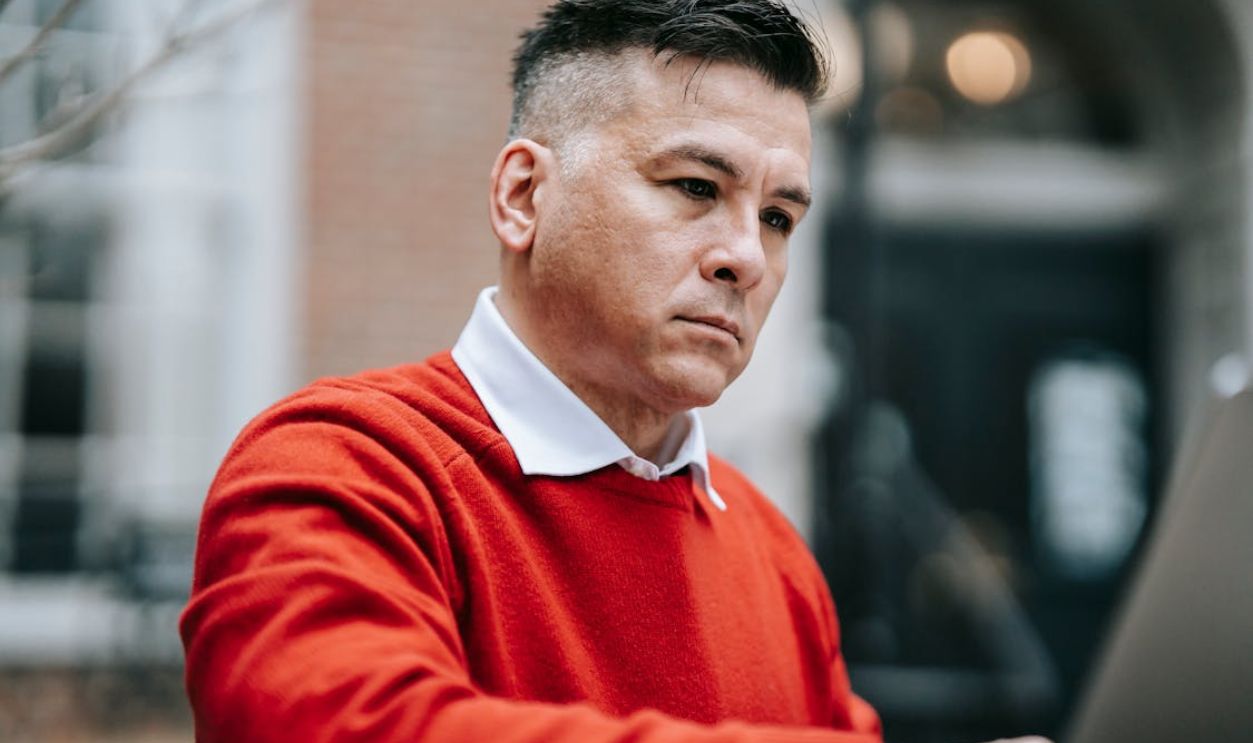 Serious man in red sweater focused on work using laptop outside an office building