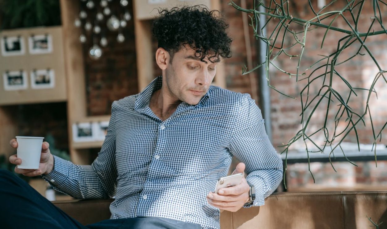 Man in stylish attire enjoying coffee while checking phone in a trendy cafe setting