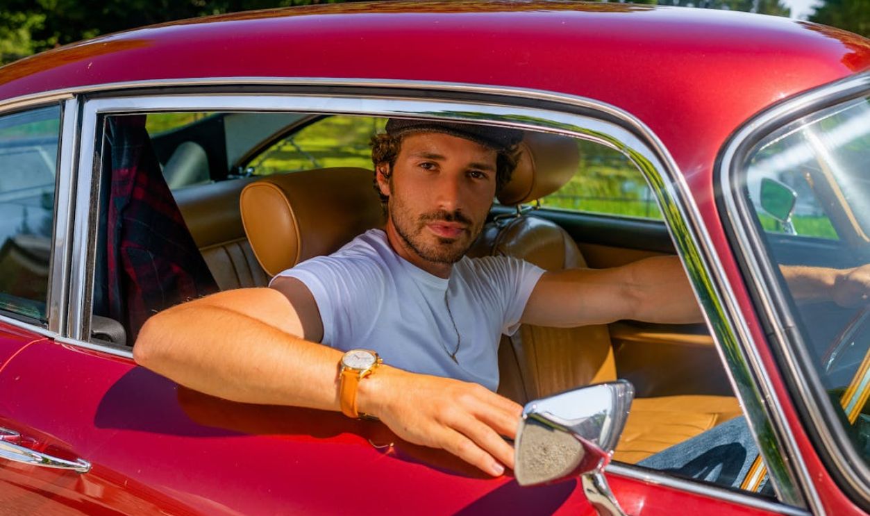 A stylish man in a white shirt drives a vintage red convertible on a sunny day