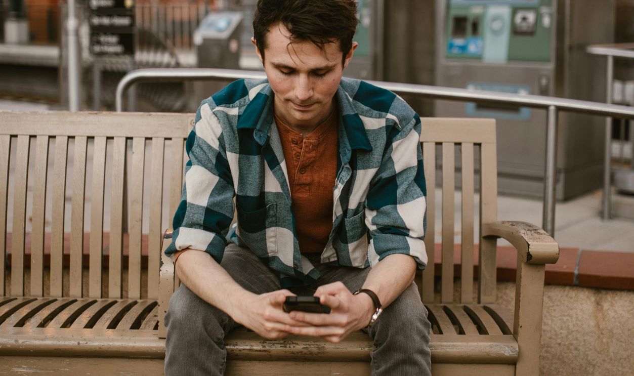 Man wearing plaid shirt using smartphone on a wooden bench at an urban train station