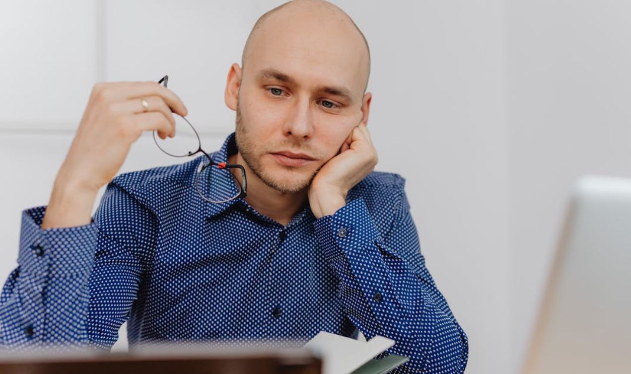 Bald businessman in blue shirt thinking at desk with glasses and laptop.