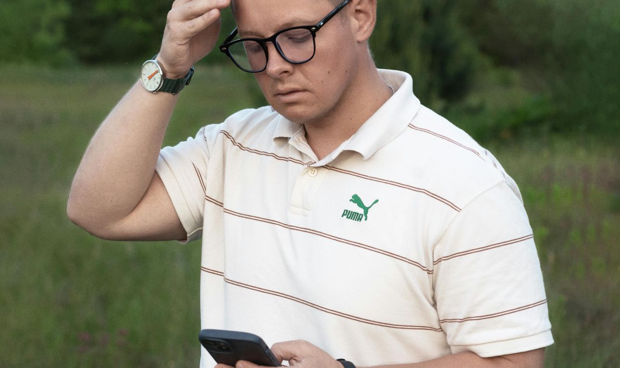 A young man in casual wear checking his smartphone in a natural outdoor setting, looking thoughtful