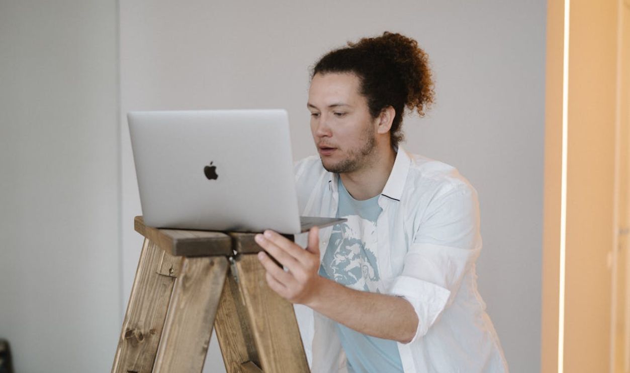 A man using a laptop while standing on a wooden ladder in a minimal indoor setting